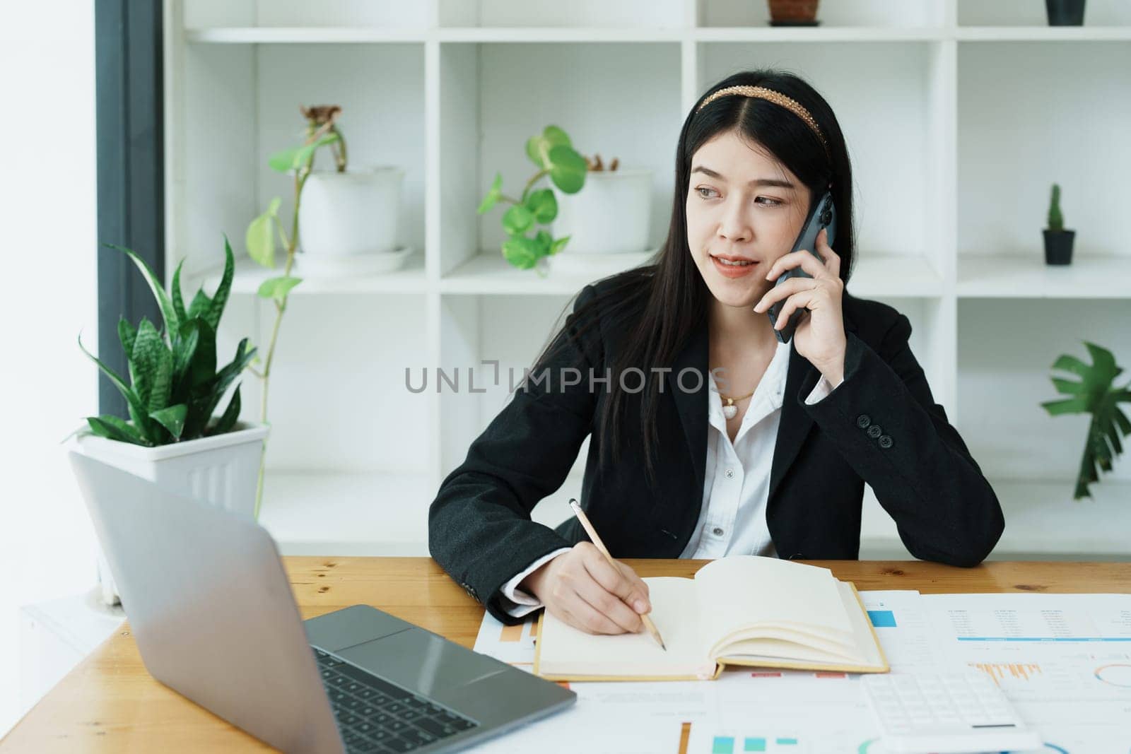 Portrait of a young Asian woman showing a smiling face as she uses her phone, computer and financial documents on her desk in the early morning hours.