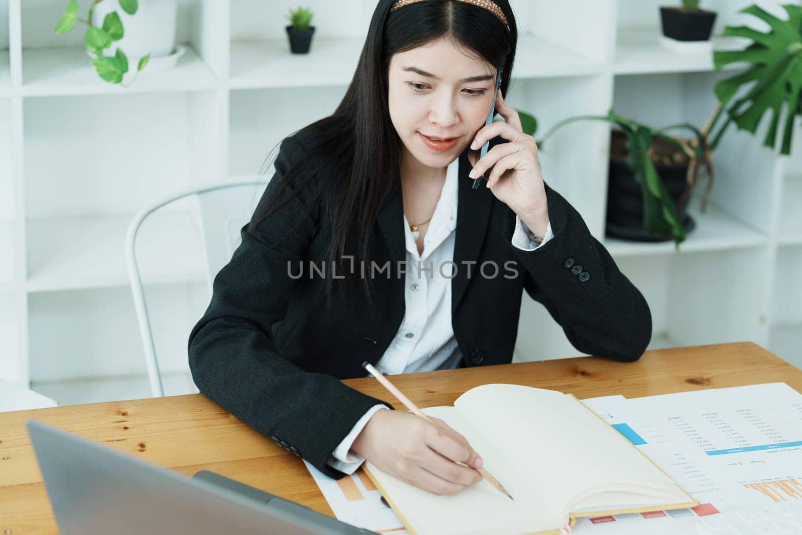 Portrait of a young Asian woman showing a smiling face as she uses her phone, computer and financial documents on her desk in the early morning hours by Manastrong