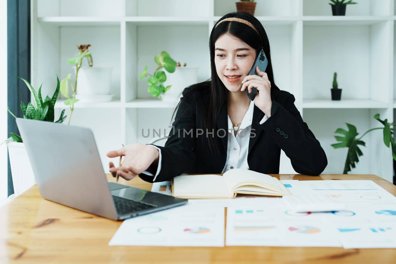 Portrait of a young Asian woman showing a smiling face as she uses her phone, computer and financial documents on her desk in the early morning hours by Manastrong