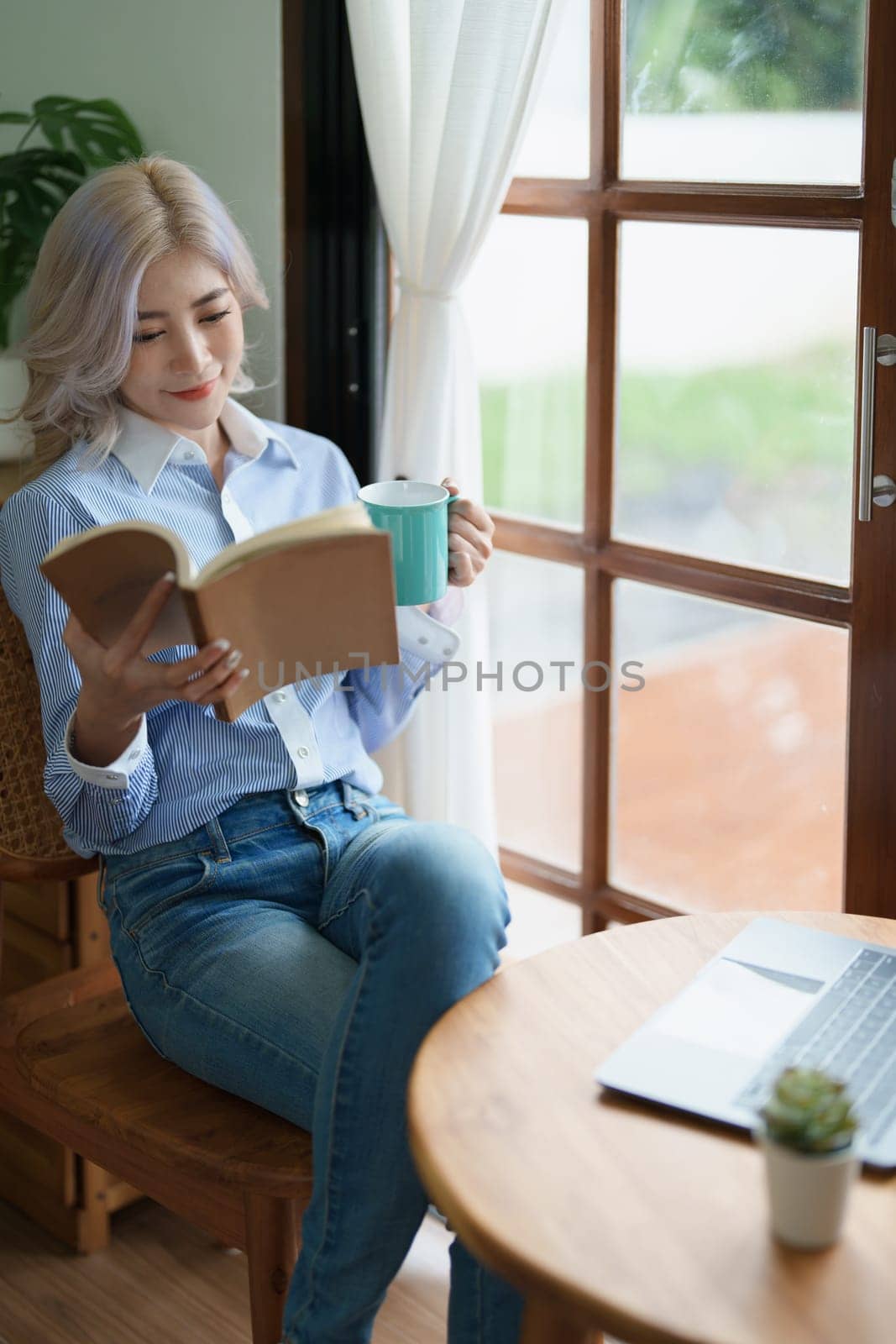 Portrait of an Asian business woman drinking coffee while reading book with a computer on her desk.