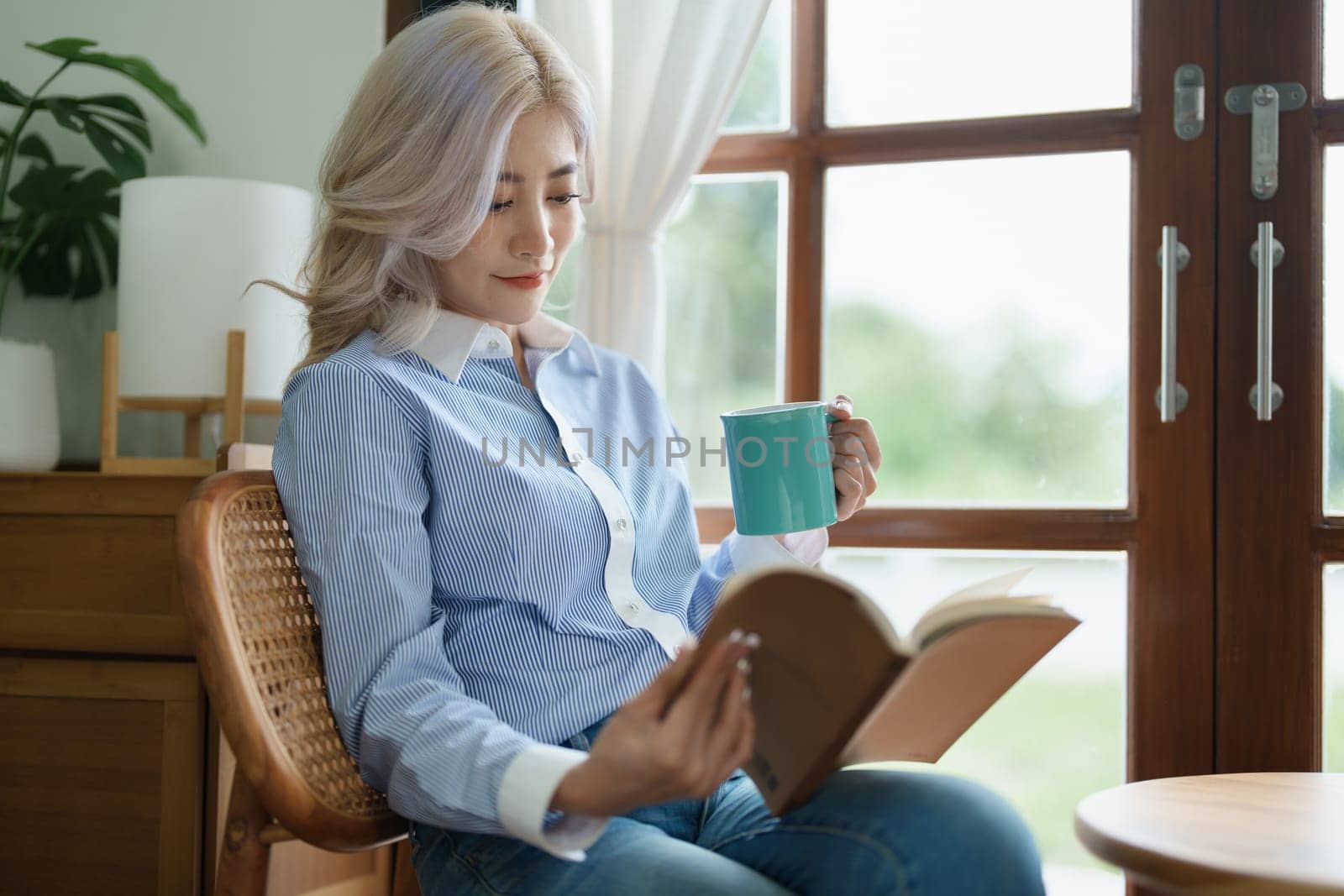 Portrait of an Asian business woman drinking coffee while reading book with a computer on her desk.