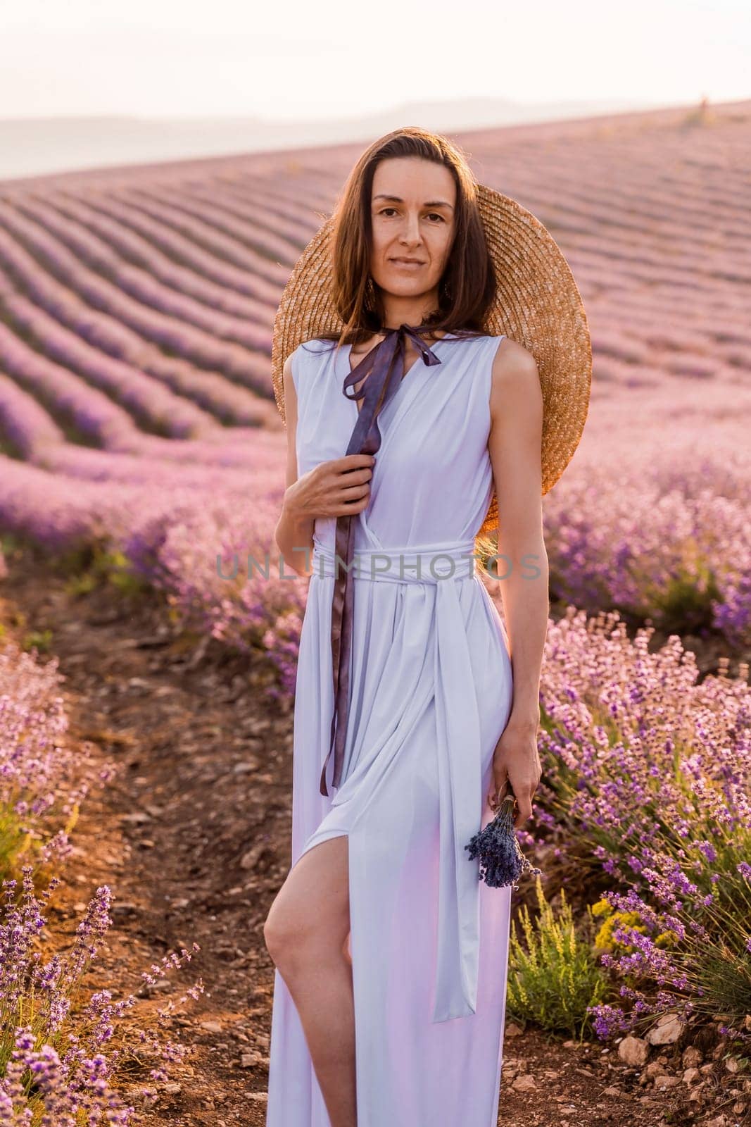 Woman lavender field sunset. Romantic woman walks through the lavender fields. illuminated by sunset sunlight. She is dressed in a white dress with a hat
