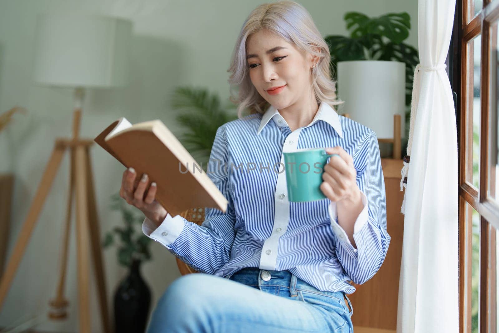 Portrait of an Asian business woman drinking coffee while reading book with a computer on her desk.