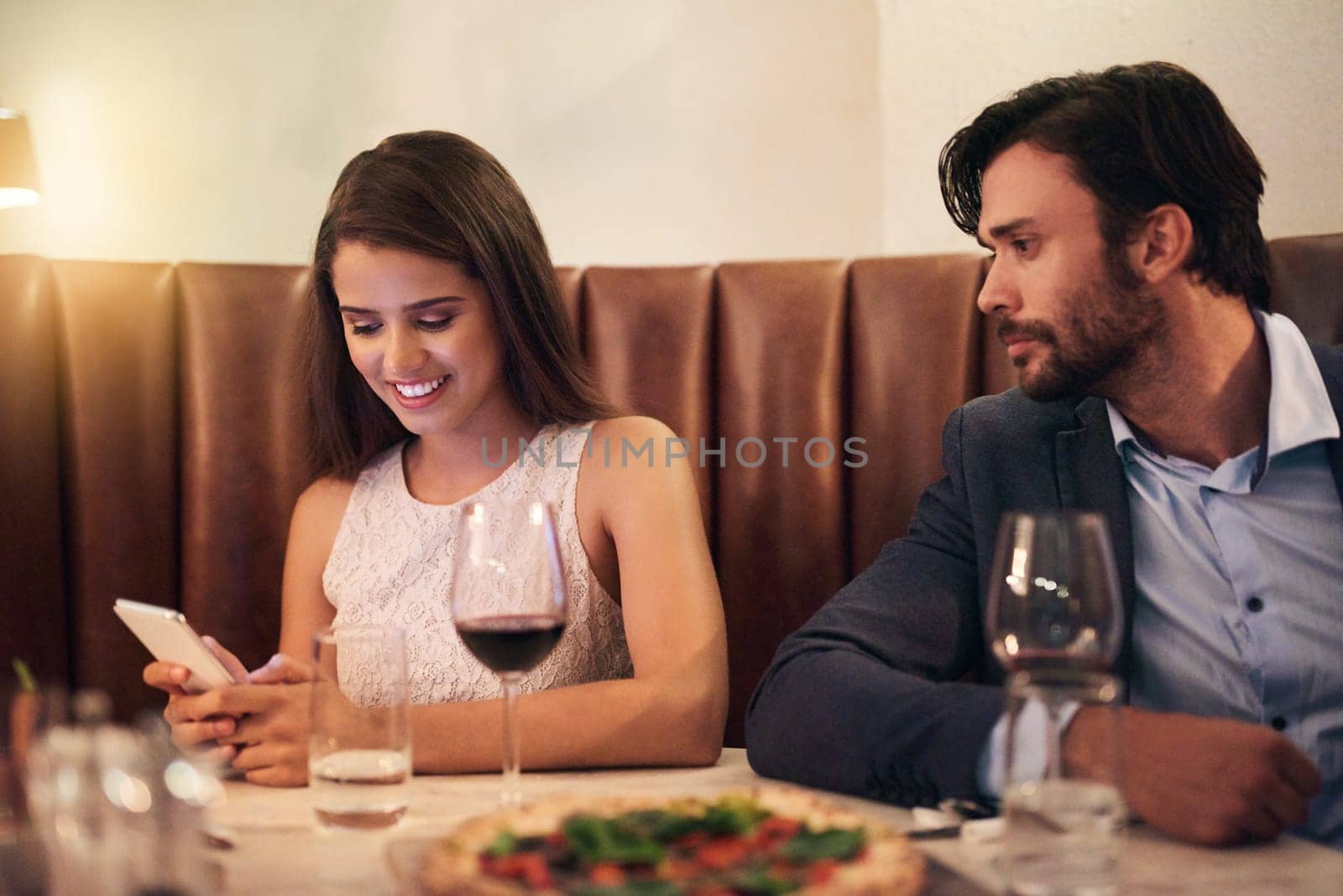 Cellphone, chatting and couple on a date at a restaurant for valentines day, romance or anniversary. Communication, upset and annoyed man watching his happy girlfriend sitting on her phone at dinner