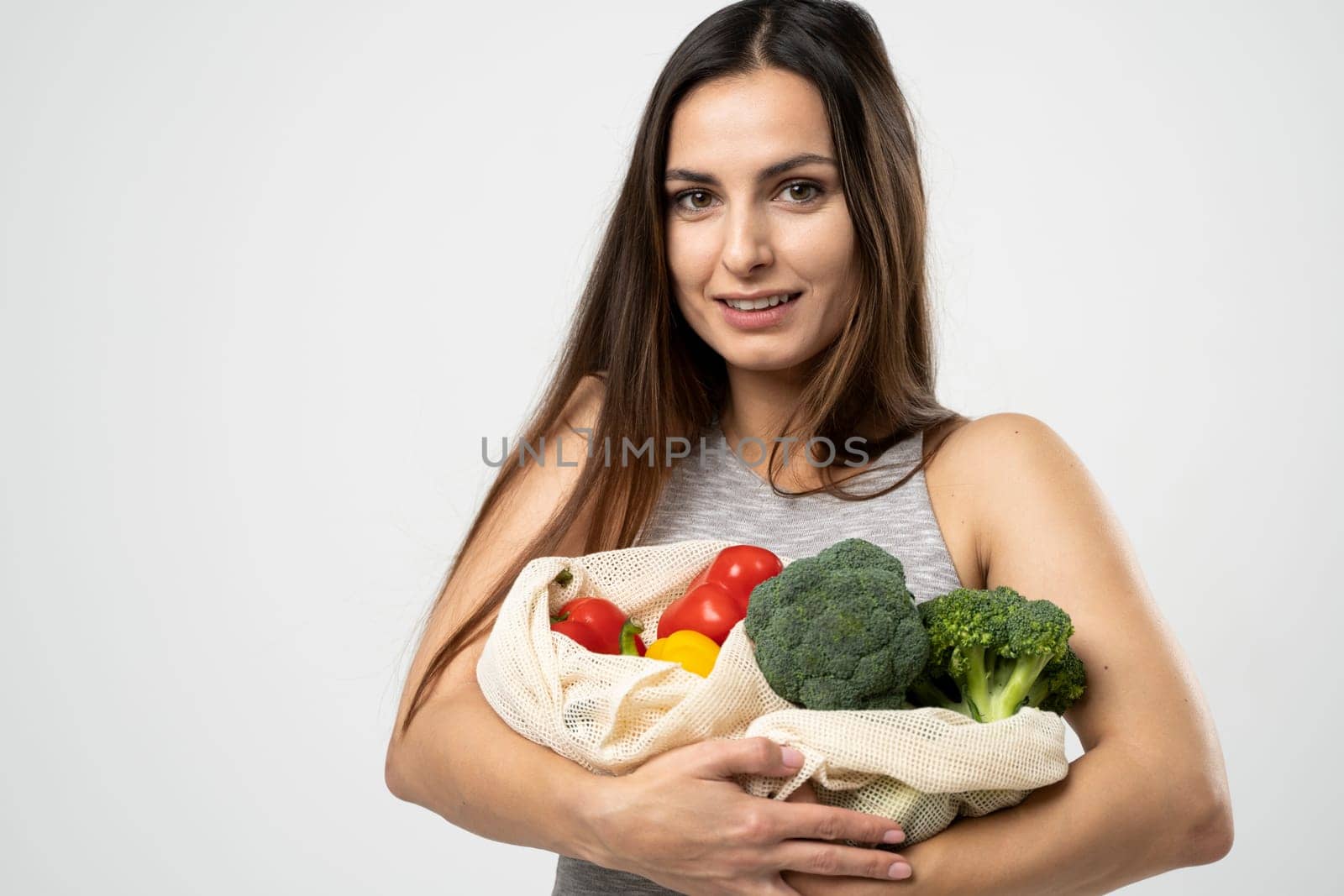 Happy smiling woman in grey dress holding reusable net bag with fruits and vegetables on white background. Food shopping and eco friendly concept. by vovsht