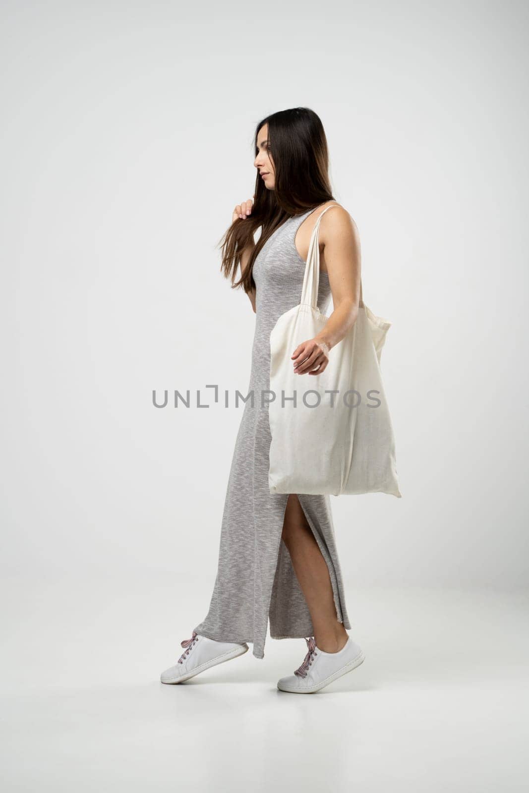 Brunette woman in grey dress holding cotton shopper bag with vegetables, products in white room. Eco friendly shopping bags. Zero waste, plastic free concept