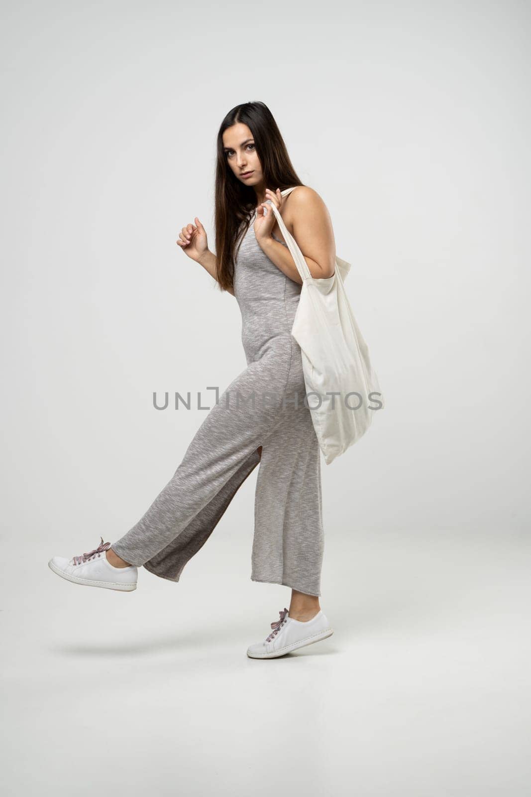 Brunette woman in grey dress holding cotton shopper bag with vegetables, products in white room. Eco friendly shopping bags. Zero waste, plastic free concept