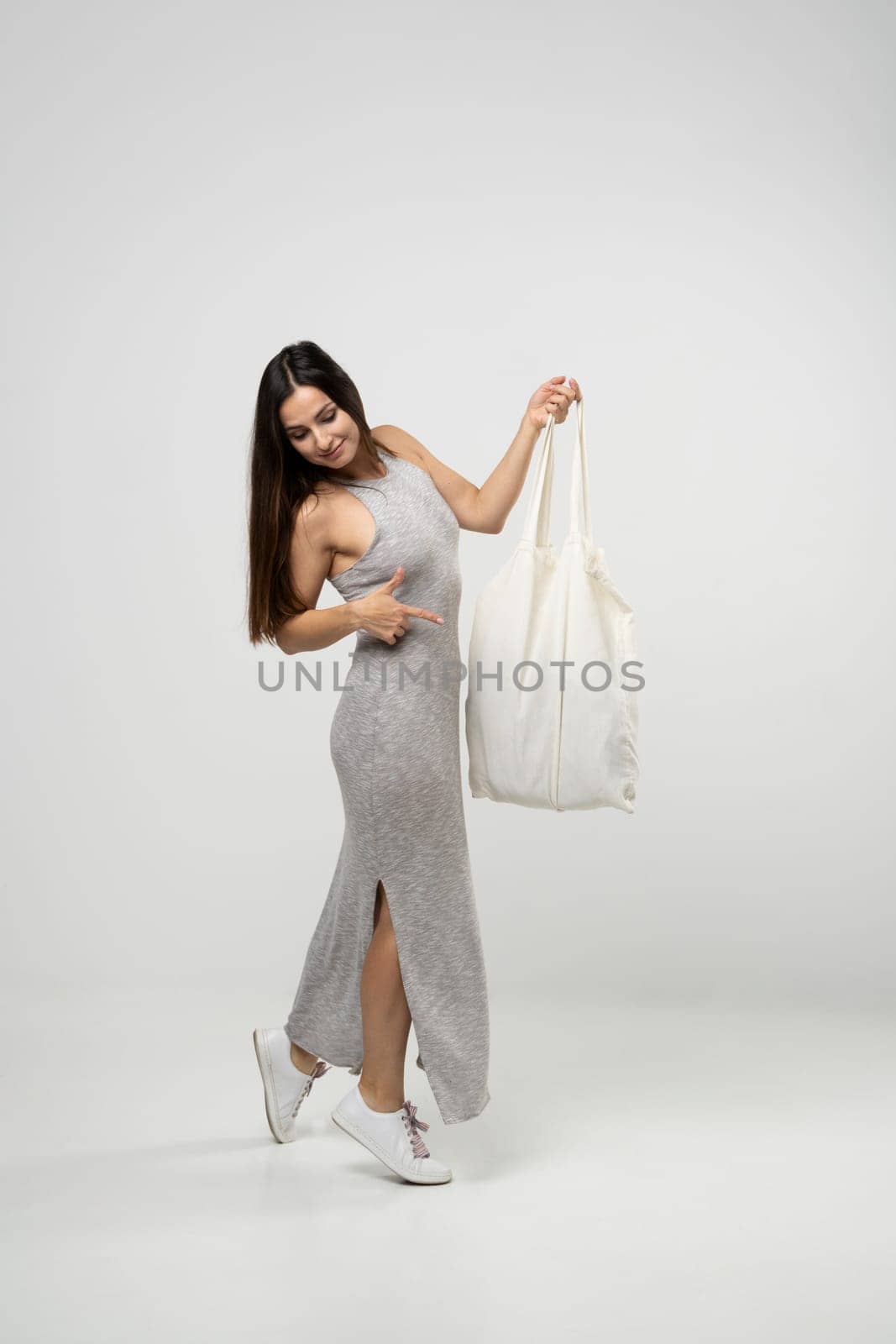 Happy young brunette woman in a grey dress dancing with a cotton bag with groceries. Zero waste concept. Eco friendly lifestyle. Mockup