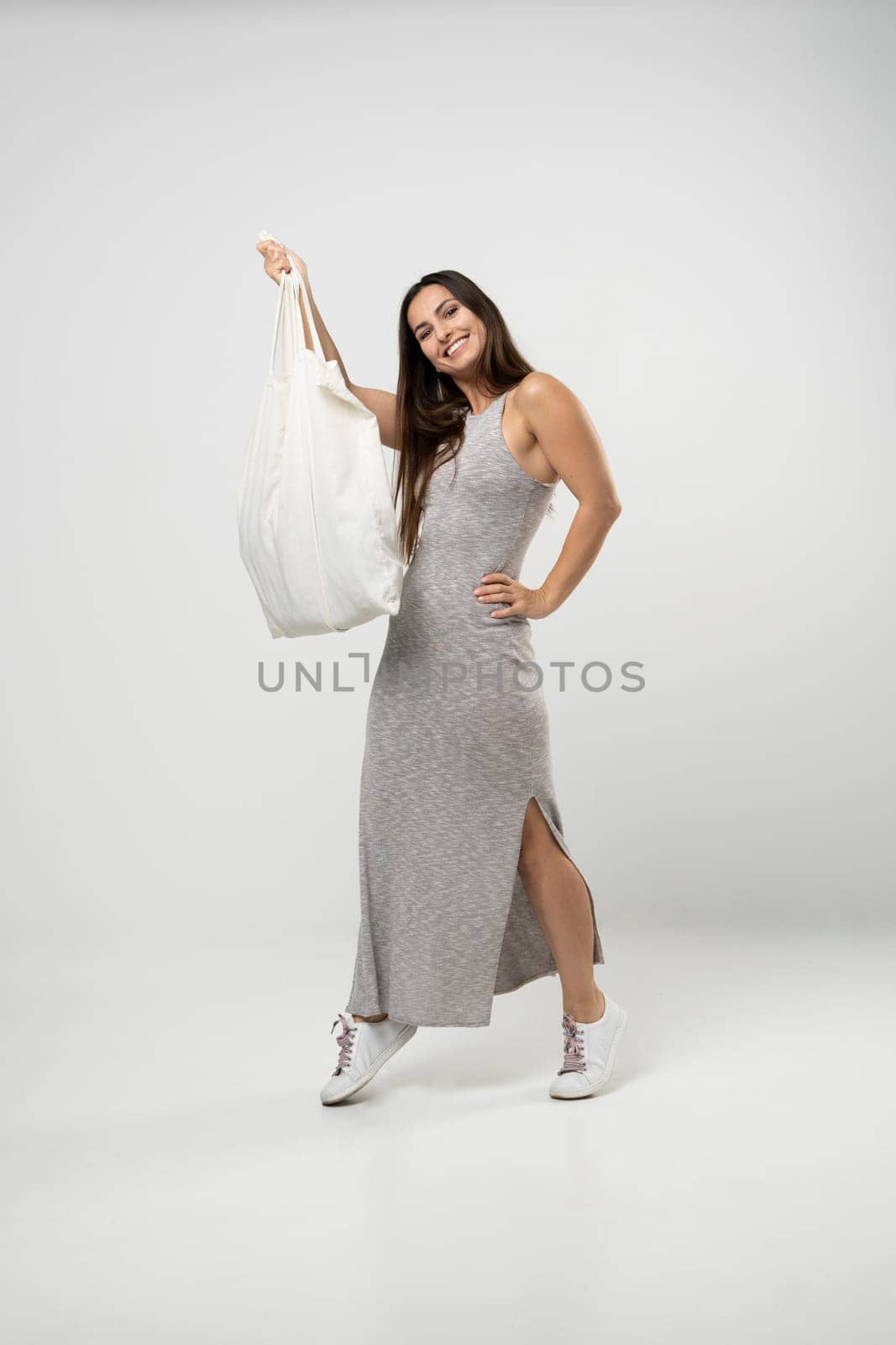 Brunette woman in grey dress holding cotton shopper bag with vegetables, products in white room. Eco friendly shopping bags. Zero waste, plastic free concept