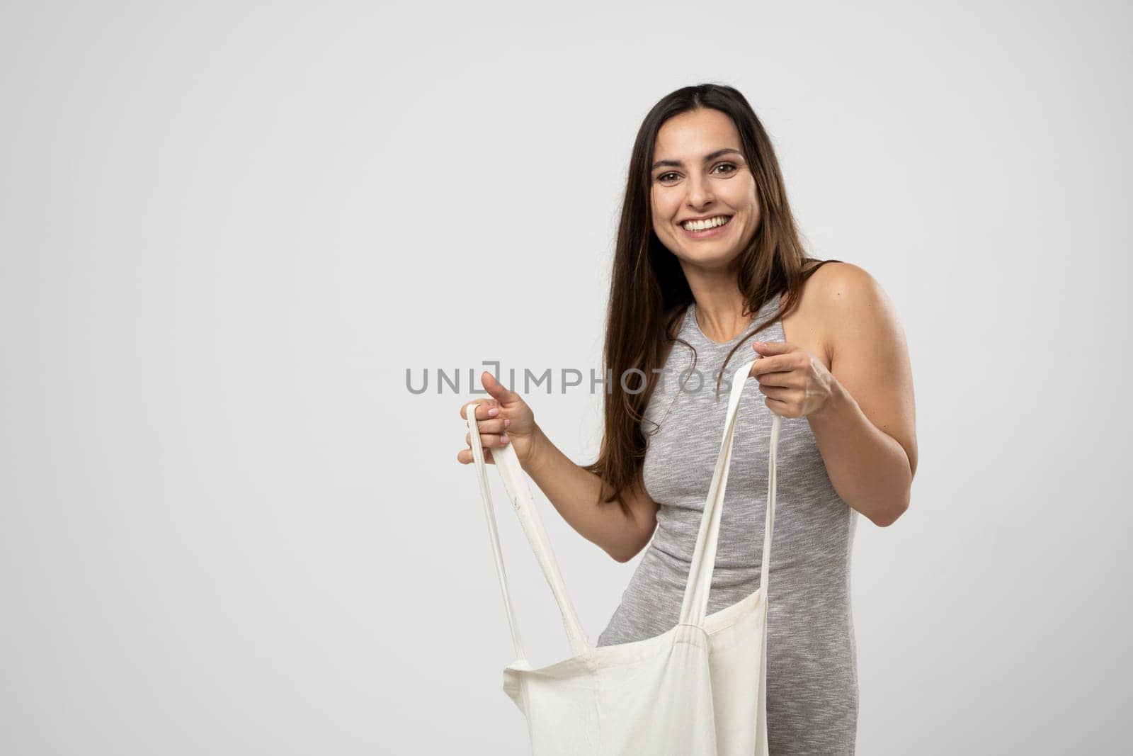 Cheerful millennial woman showing white eco bag to camera standing over white studio background. Lady holding cotton shopper handbag. fashion and ecology concept. by vovsht