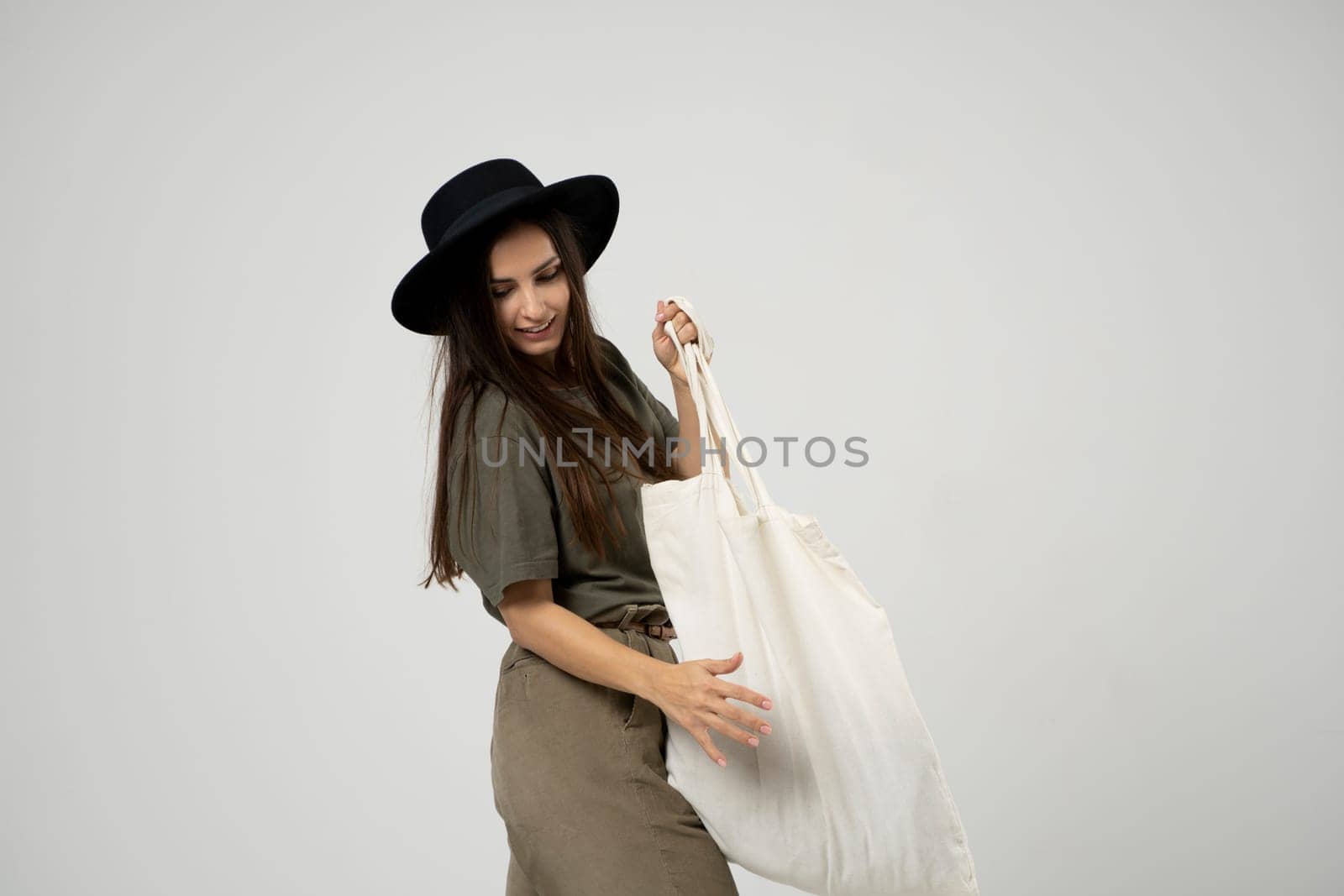 Brunette woman in green t-shirt and black hat holding cotton shopper bag with vegetables, products in white room. Eco friendly shopping bags. Zero waste, plastic free concept. by vovsht