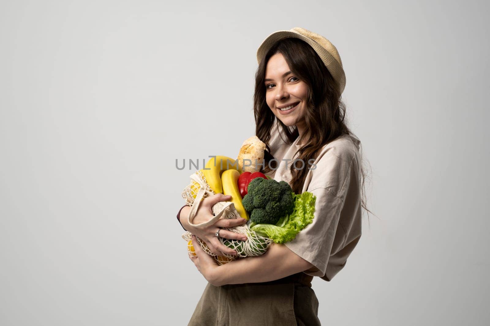 Zero waste concept. Brunette woman in a beige t-shirt and a hat with a mesh bag full of fresh vegetables and fruits after supermarket. Sustainable lifestyle with no plastic