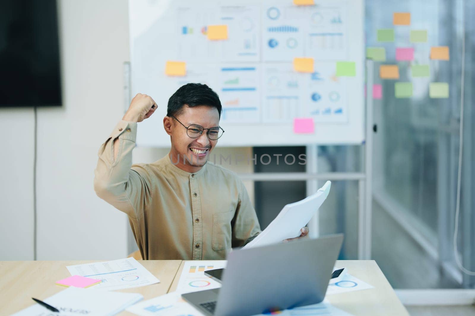 Portrait of a male business owner showing a happy smiling face as he has successfully invested his business using computers and financial budget documents at work.