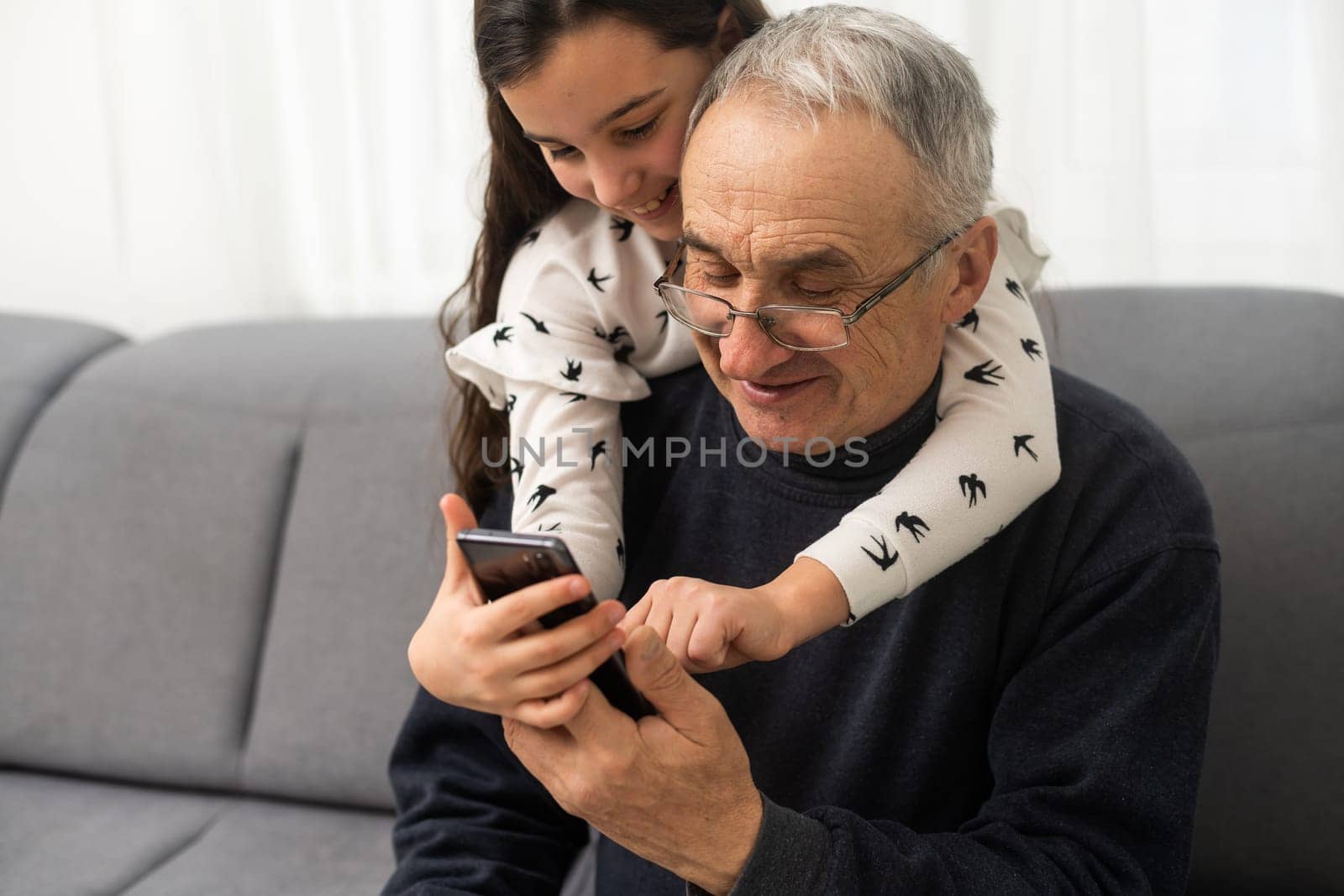 senior grandfather learning to using mobile phone under guidance of pretty young granddaughter sitting on sofa at home by Andelov13