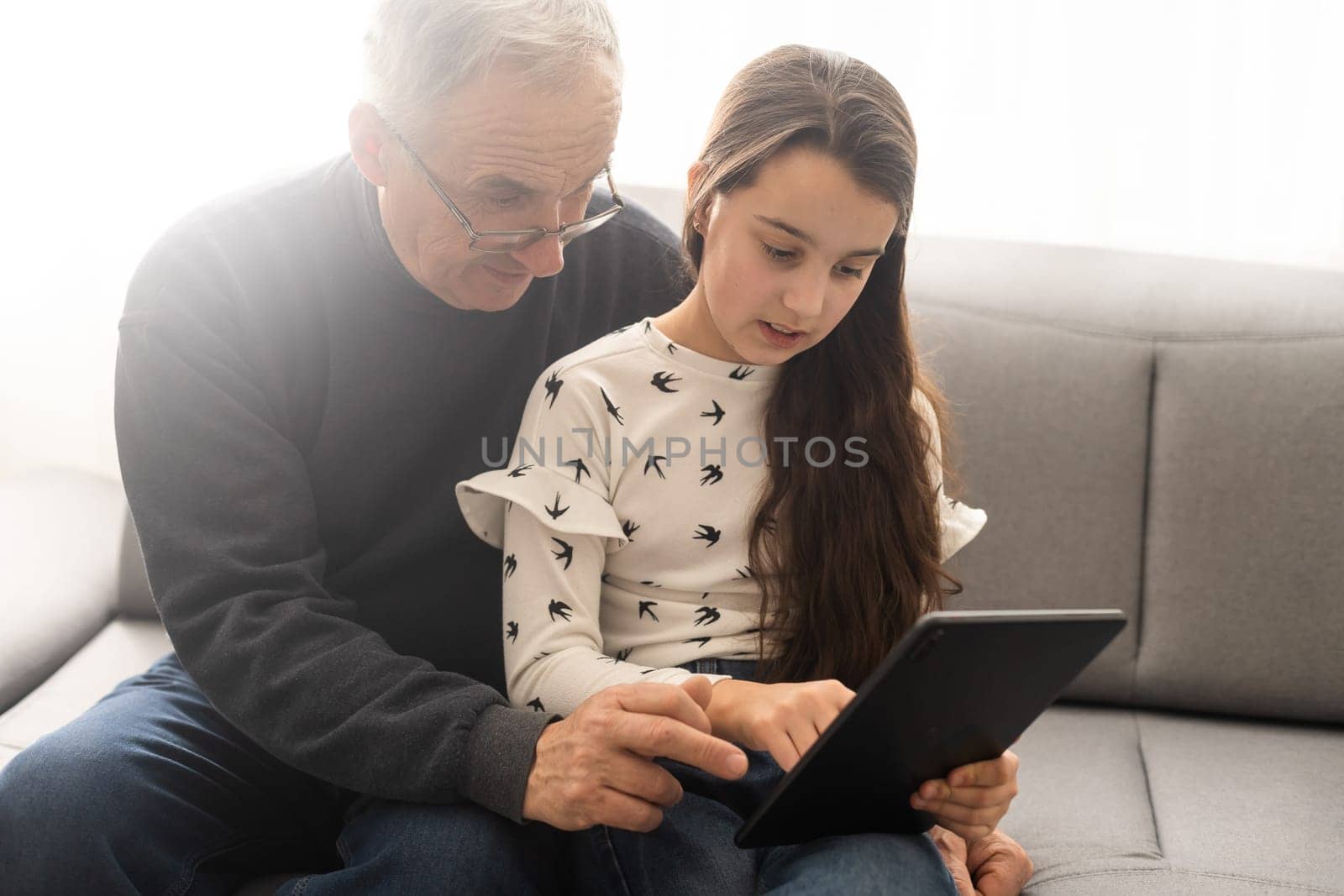 Cute little girl and her handsome grandpa are smiling while sitting on couch at home. Girl is using a tablet. by Andelov13