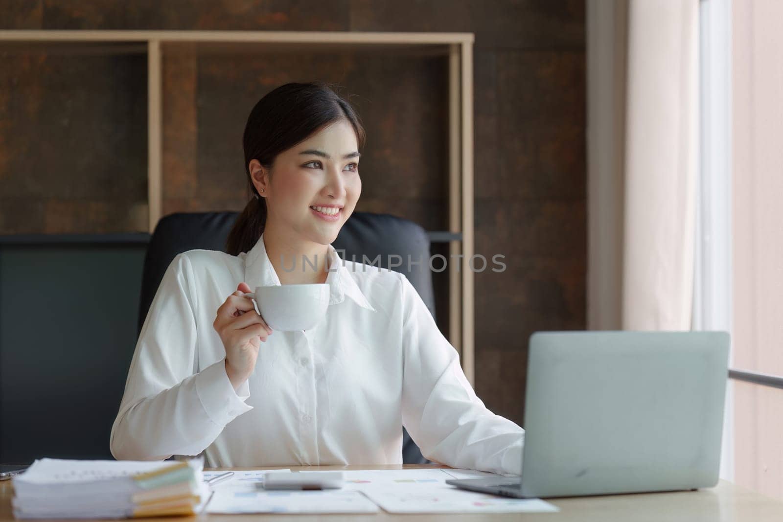 Attractive asian businesswoman sitting and drinking coffee at the desk in home office.