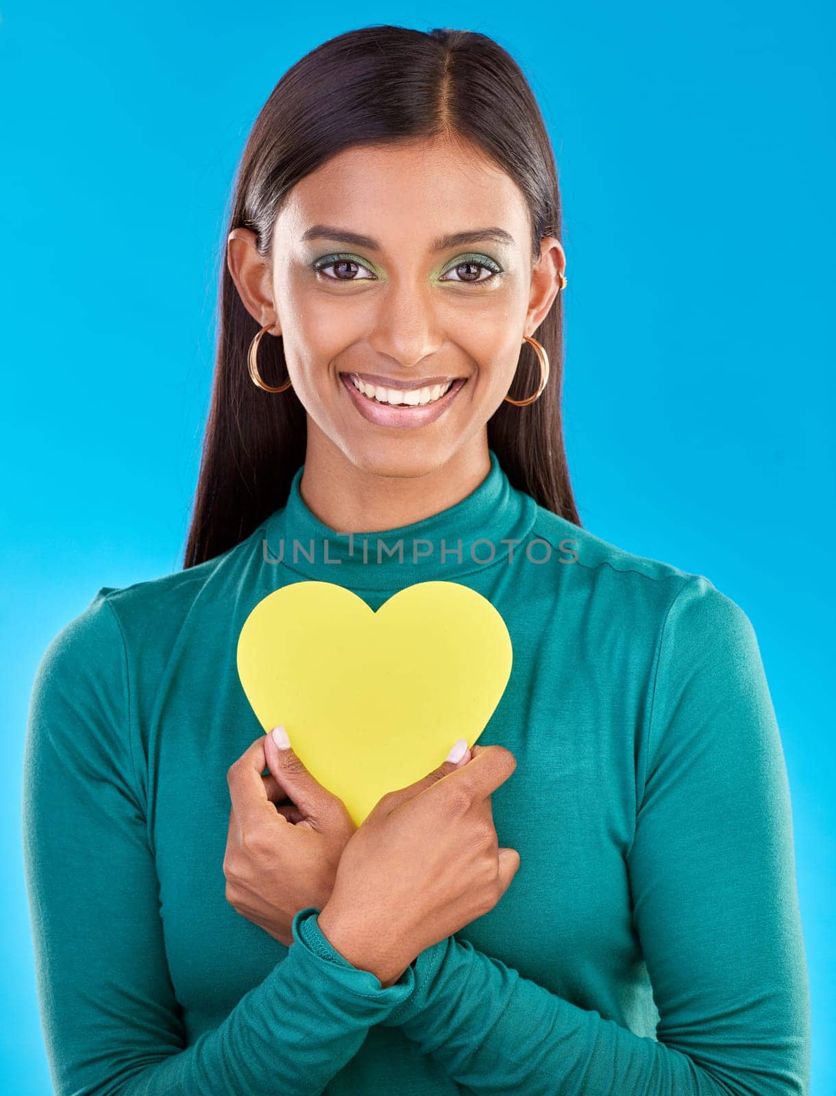 Portrait, makeup and heart on valentines day with a woman in studio on a blue background for love or health. Face, beauty and romance with an attractive young female holding a cut out emoji or symbol by YuriArcurs
