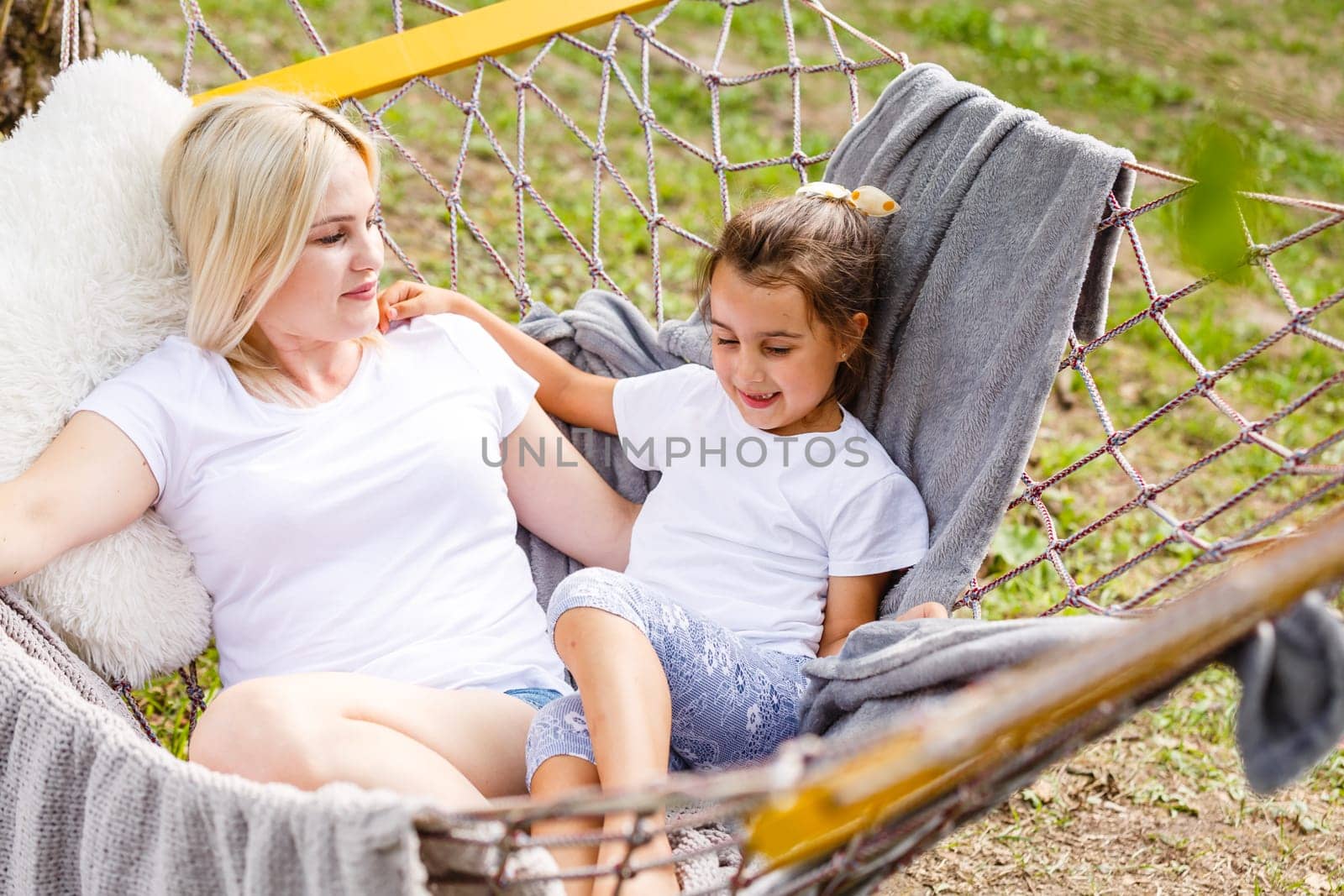 Wide view of a young mother and daughter relaxing together and smiling sitting in a hammock, hugging and lounging during a sunny summer day in a holiday home garden with grass and trees, lifestyle.