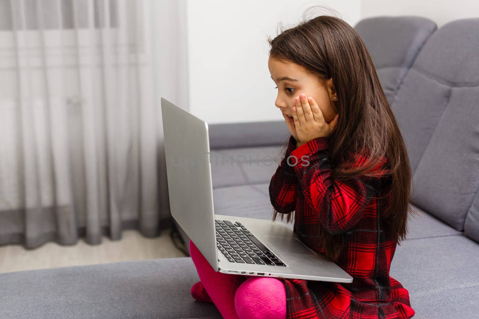 The little girl sits at a table with surprise looks at the laptop. On a white background.