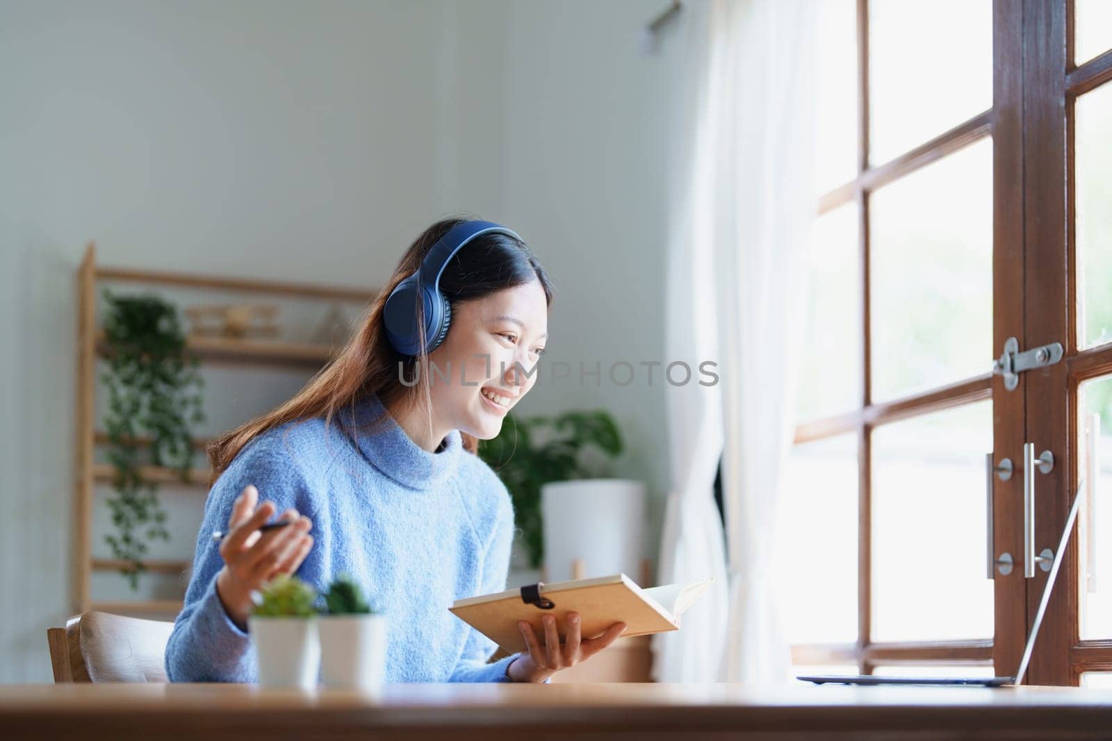 Portrait of a teenage Asian woman using a computer, wearing headphones and using a notebook to study online via video conferencing on a wooden desk at home by Manastrong