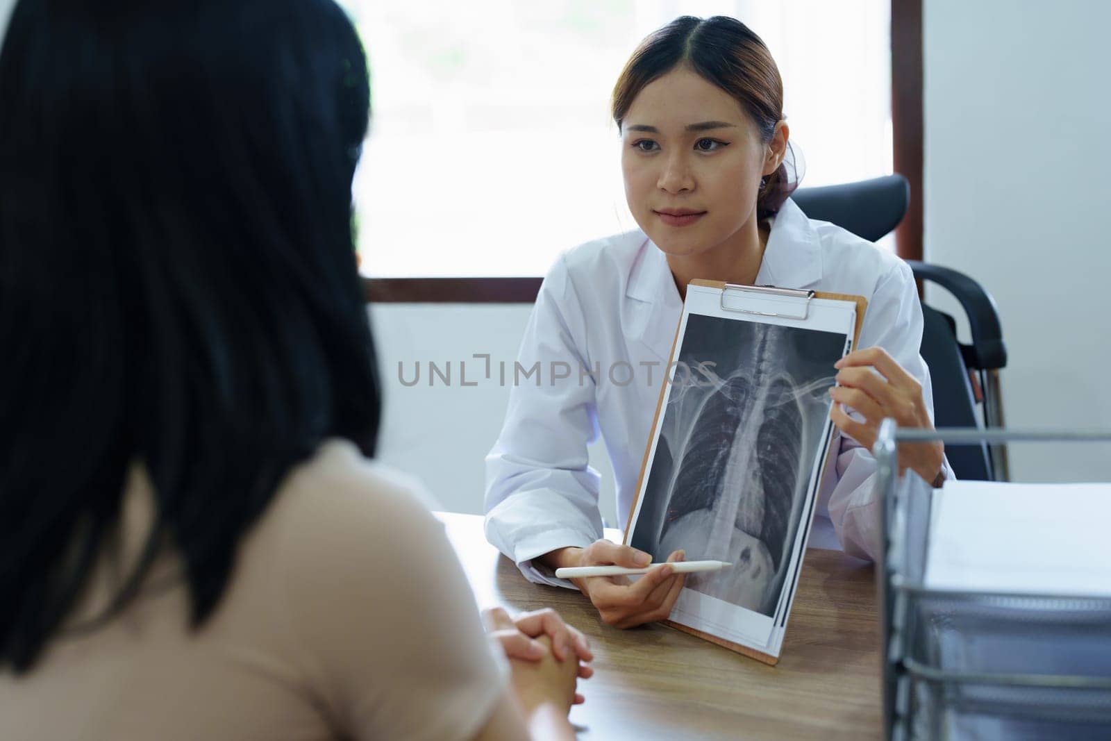 An Asian female doctor points to a patient x-ray film to explain the patient's treatment process by Manastrong