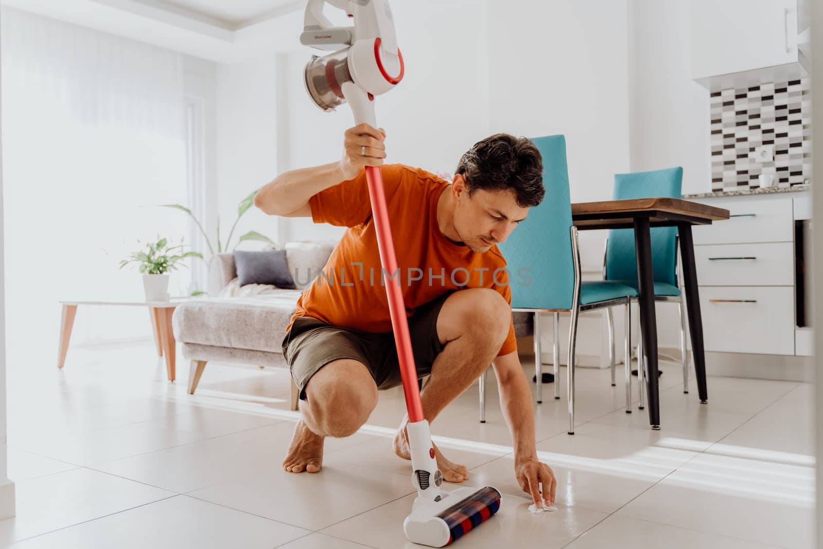 White man male sitting and wiping tiled floor in the kitchen living room with wet wipes holding vacuum cleaner brush head.