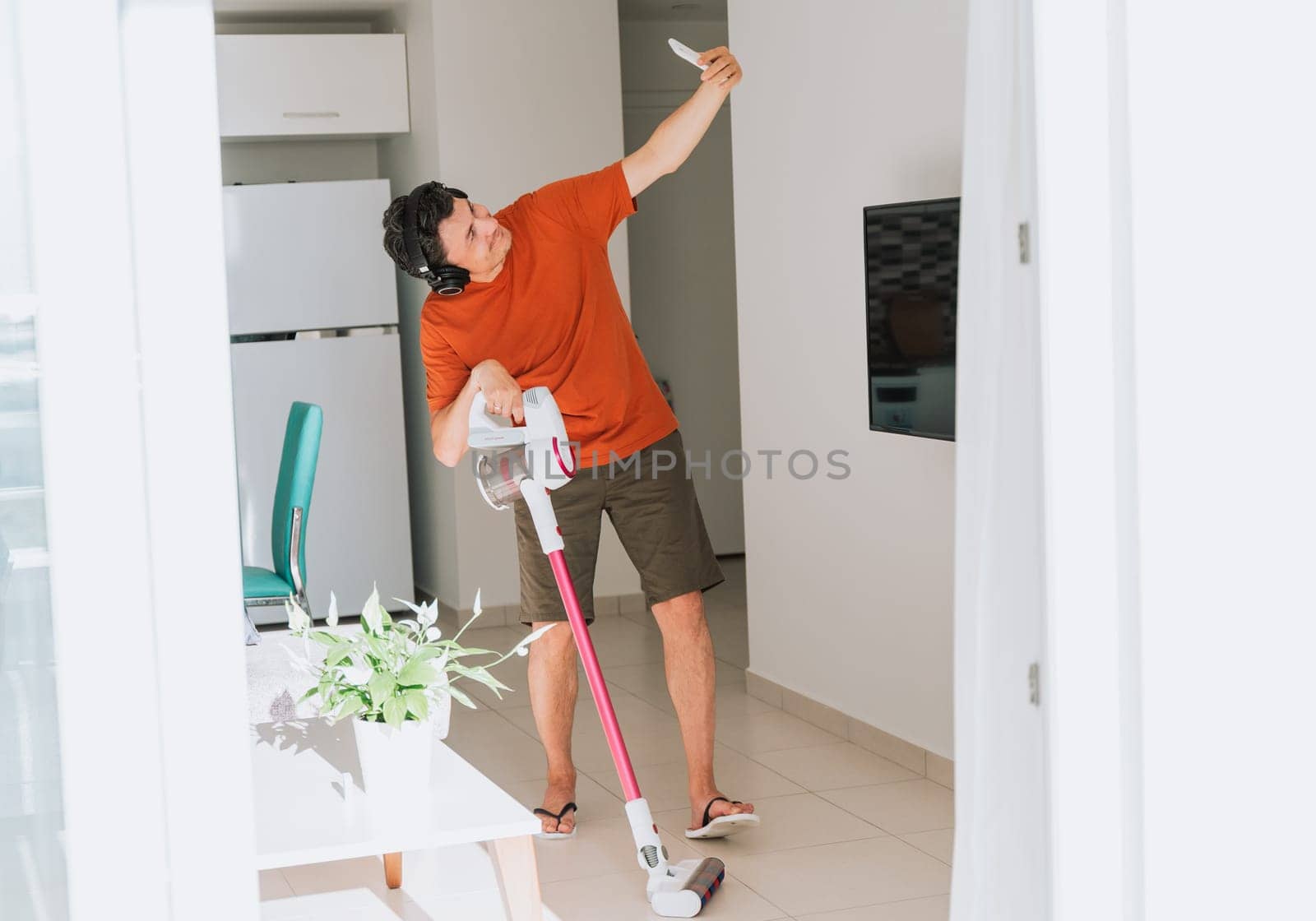 Man taking a selfie on cellphone while cleaning with wireless vacuum cleaner and headphones. White male in light living room doing home domestic chores with wireless vacuum cleaner earphones mobile