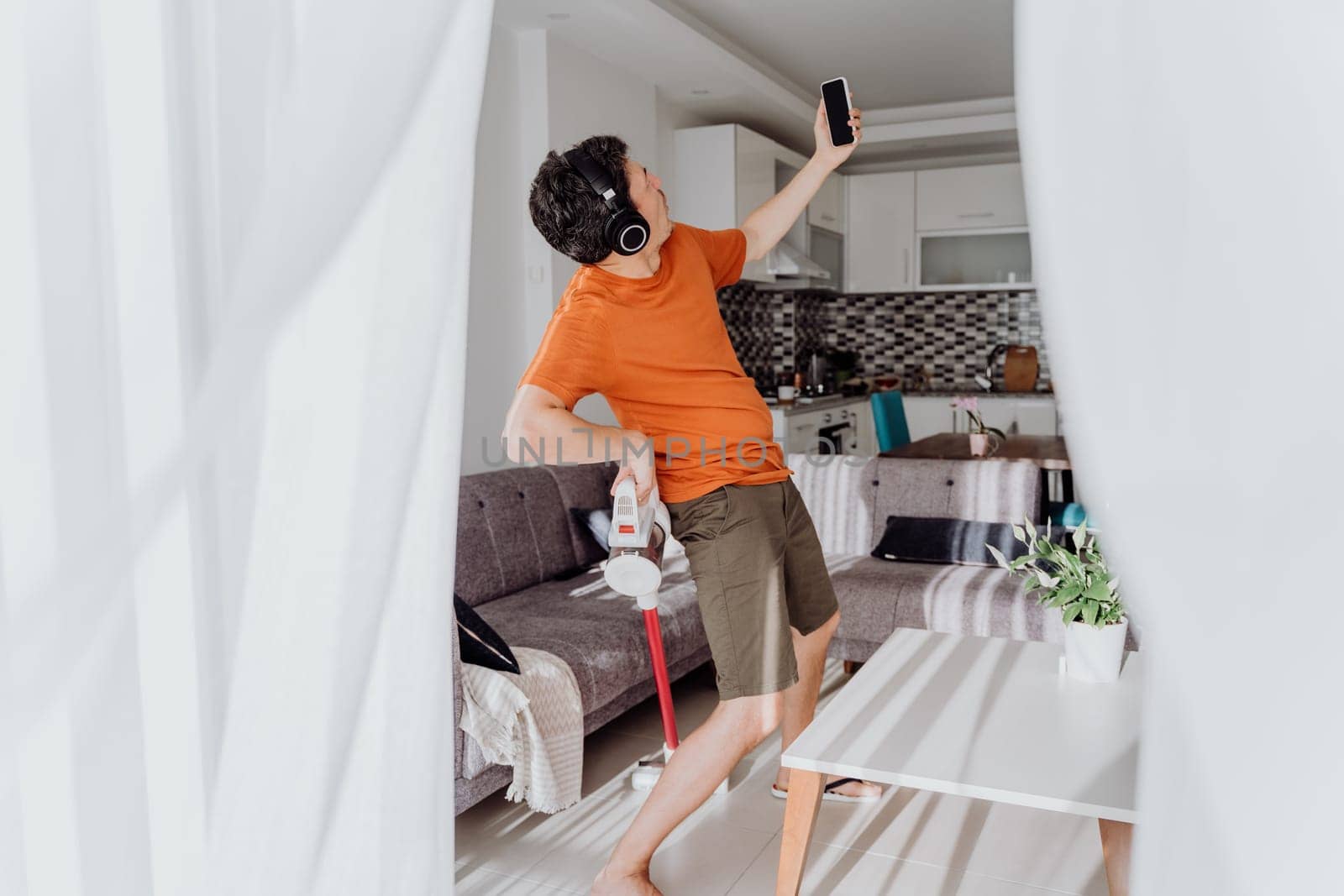 Man taking a selfie on cellphone while cleaning with wireless vacuum cleaner and headphones. White male in light living room doing home domestic chores with wireless vacuum cleaner earphones mobile