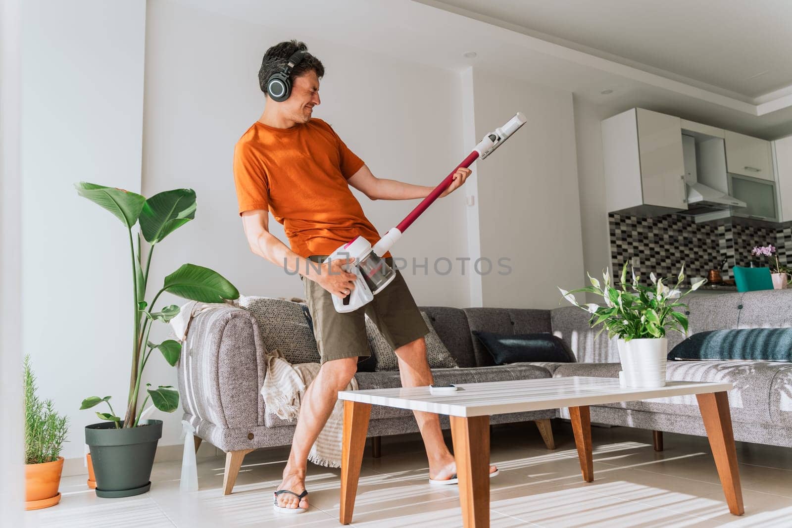 Caucasian man wearing wireless headphones plays rocks air guitar on vacuum cleaner. Male doing home chores with fun.