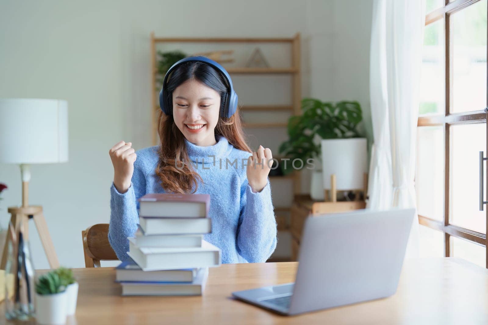 Portrait of young beautiful Asian woman showing smiling face during early morning online class with books, headphones and computer as study materials at home.