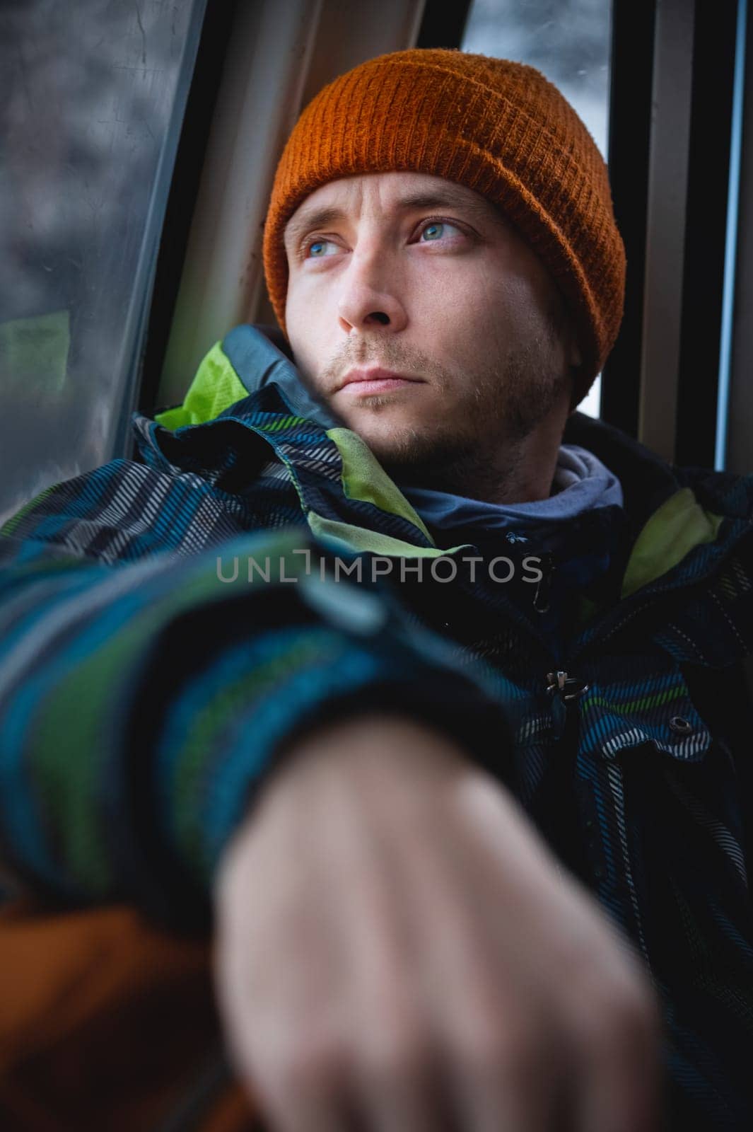 Handsome caucasian man with a beard looks thoughtfully to the side. Beautiful winter portrait of a handsome man in a knitted hat sitting in a ski cable car cabin.