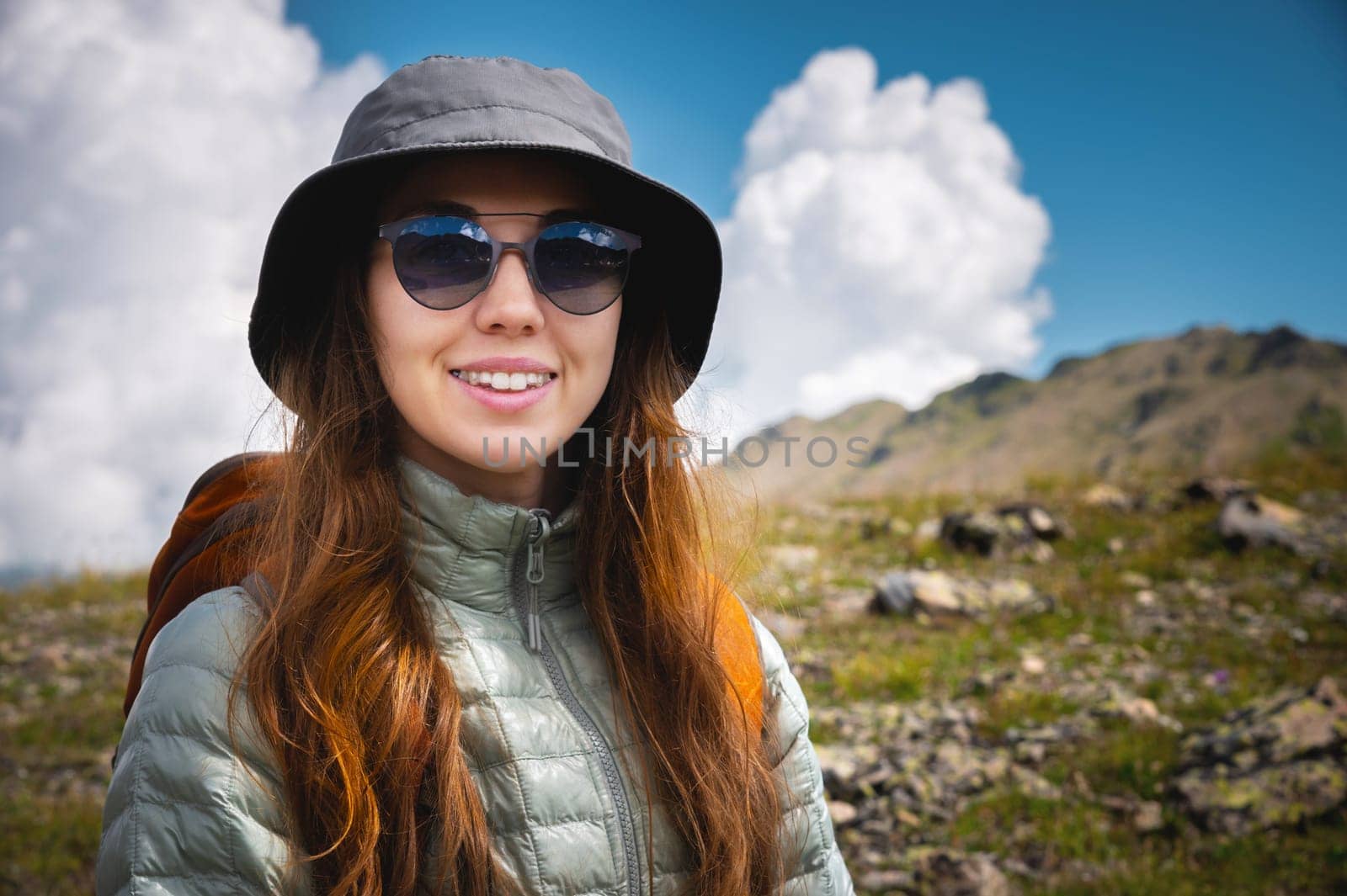 Portrait of a traveler in the mountains. The concept of adventure, travel and hiking. Happy woman in a cap enjoys the sunshine while hiking in the mountains. Tourist with glasses by yanik88