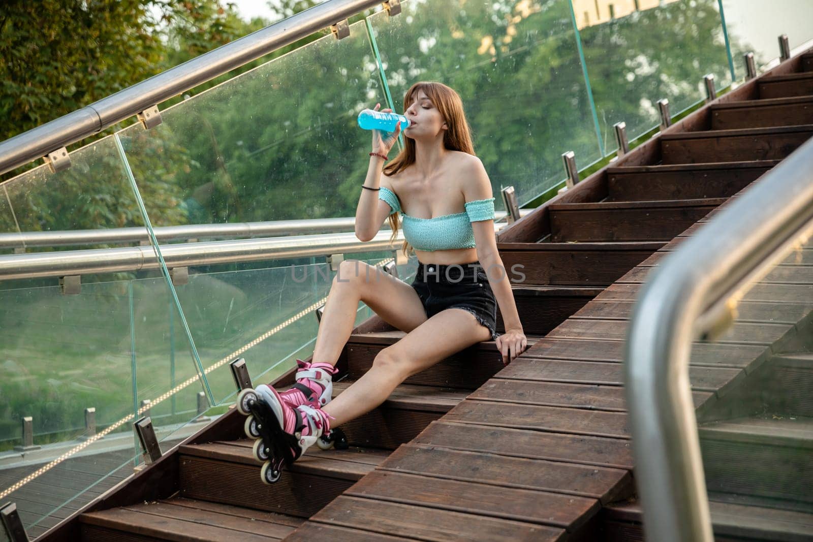 Long-haired skater girl relaxes after skating. The girl is sitting on a wooden staircase. She is drinking a from a bottle. Through the glass balustrade you can see trees and lots of greenery.