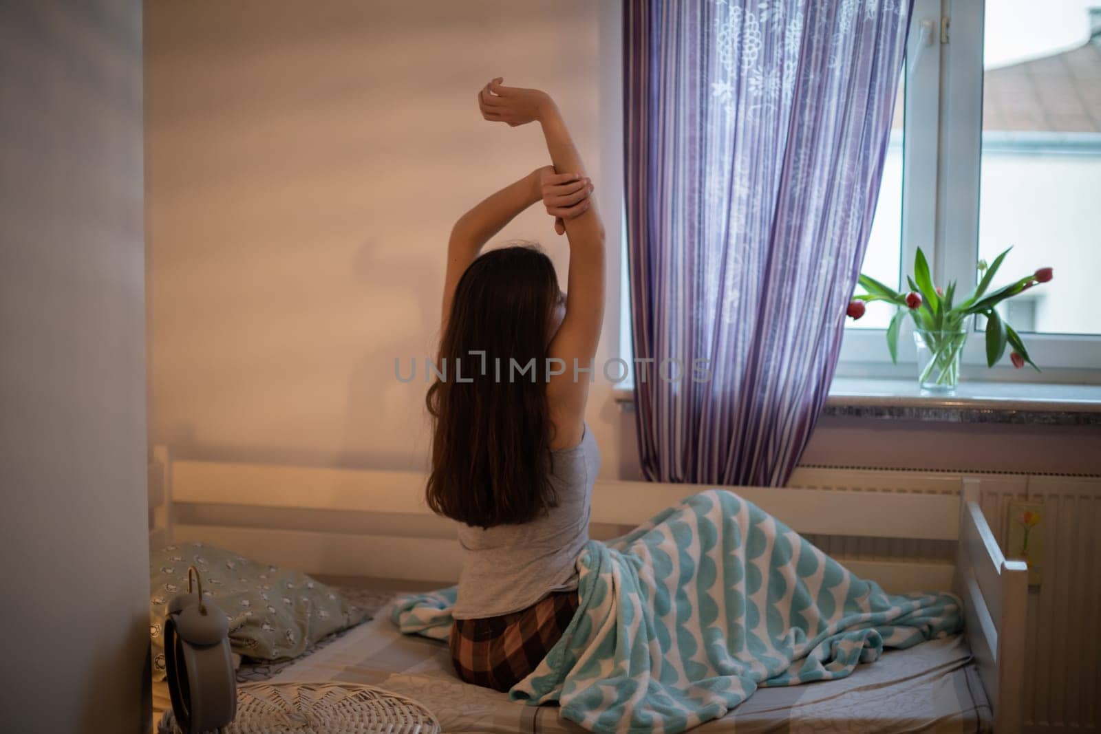 A woman with long hair drags herself and looks out the window. Bright bedroom with decorations.