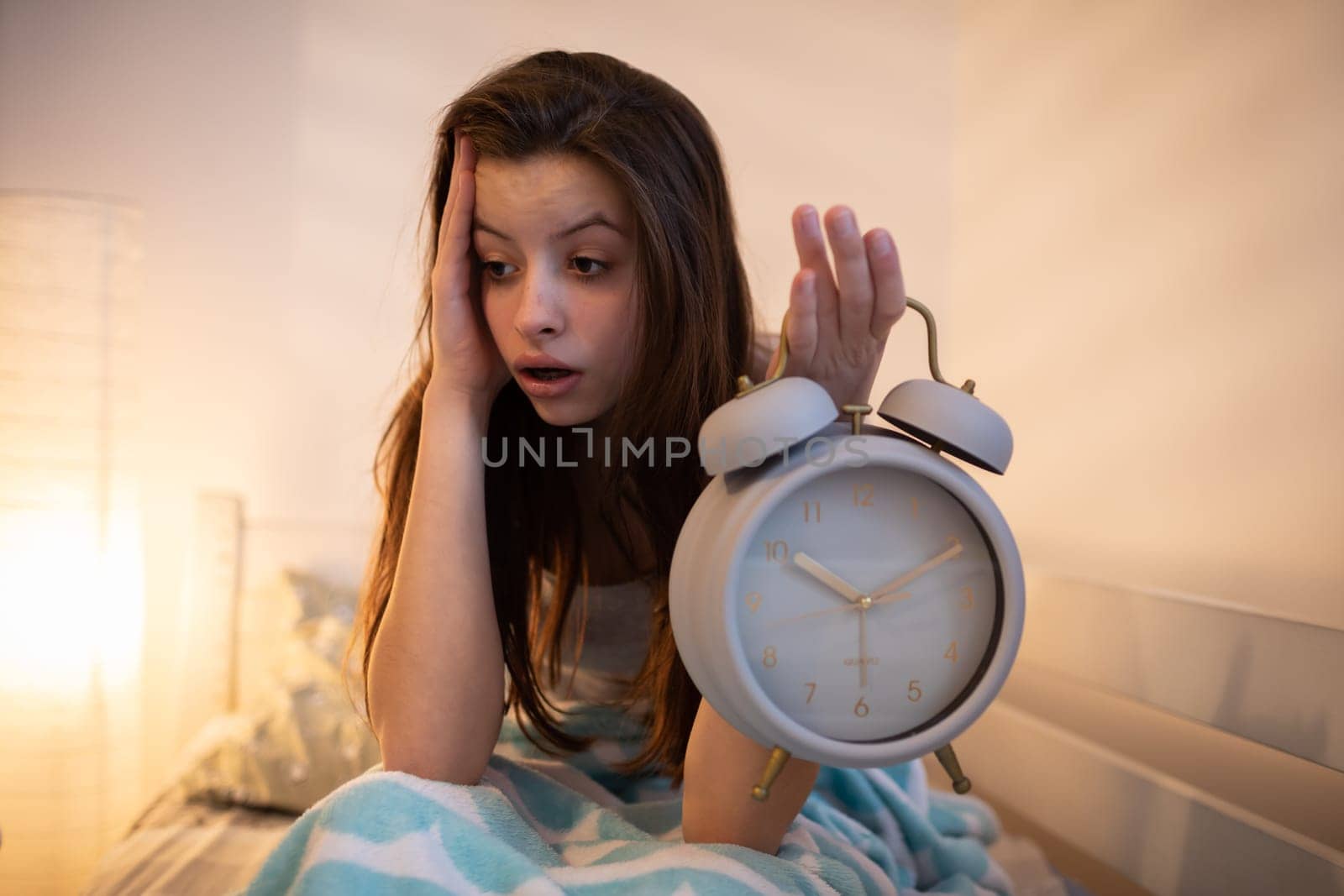 A tired girl holds a bright alarm clock in her hand. The light of a lamp on the wall.