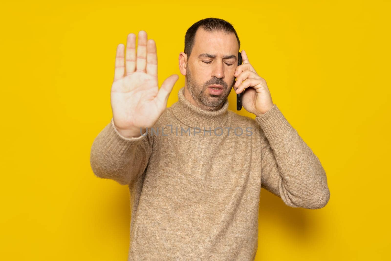 Bearded hispanic man in a turtleneck asking not to be disturbed while talking on his smartphone, he is very pissed off the interruption has taken him out of his mind. Isolated on yellow background