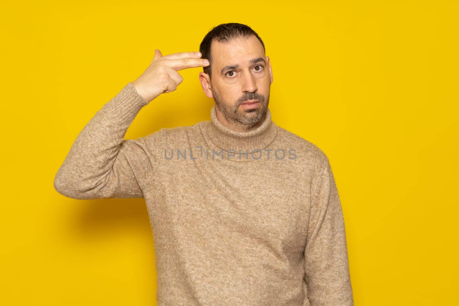 Annoyed hispanic man with beard making gun gesture with finger to head, shooting himself out of boredom or annoyance, tired of something stupid, standing over yellow background