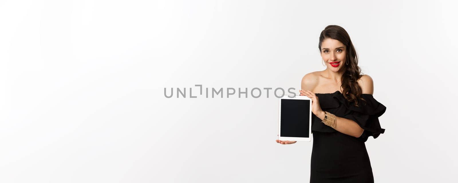 Online shopping concept. Tempted pretty woman in black dress showing digital tablet screen, standing over white background. Copy space