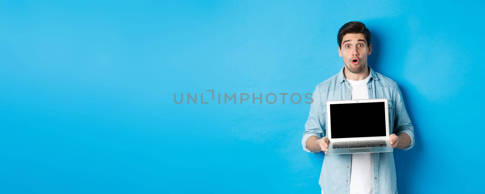 Man showing advertisement on laptop screen and looking amazed, saying wow and looking at camera, standing against blue background.