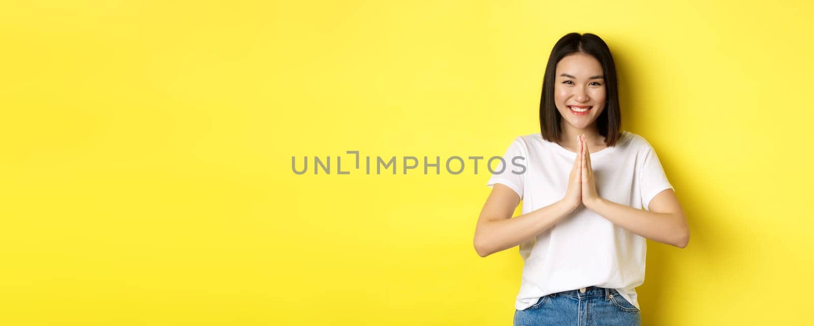 Beautiful asian woman say thank you, holding hands in namaste, pray gesture and smiling, being grateful, standing over yellow background.