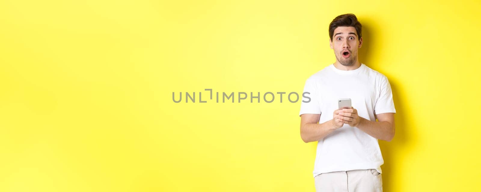 Man looking surprised, using smartphone, open mouth and saying wow, standing against yellow background. Copy space