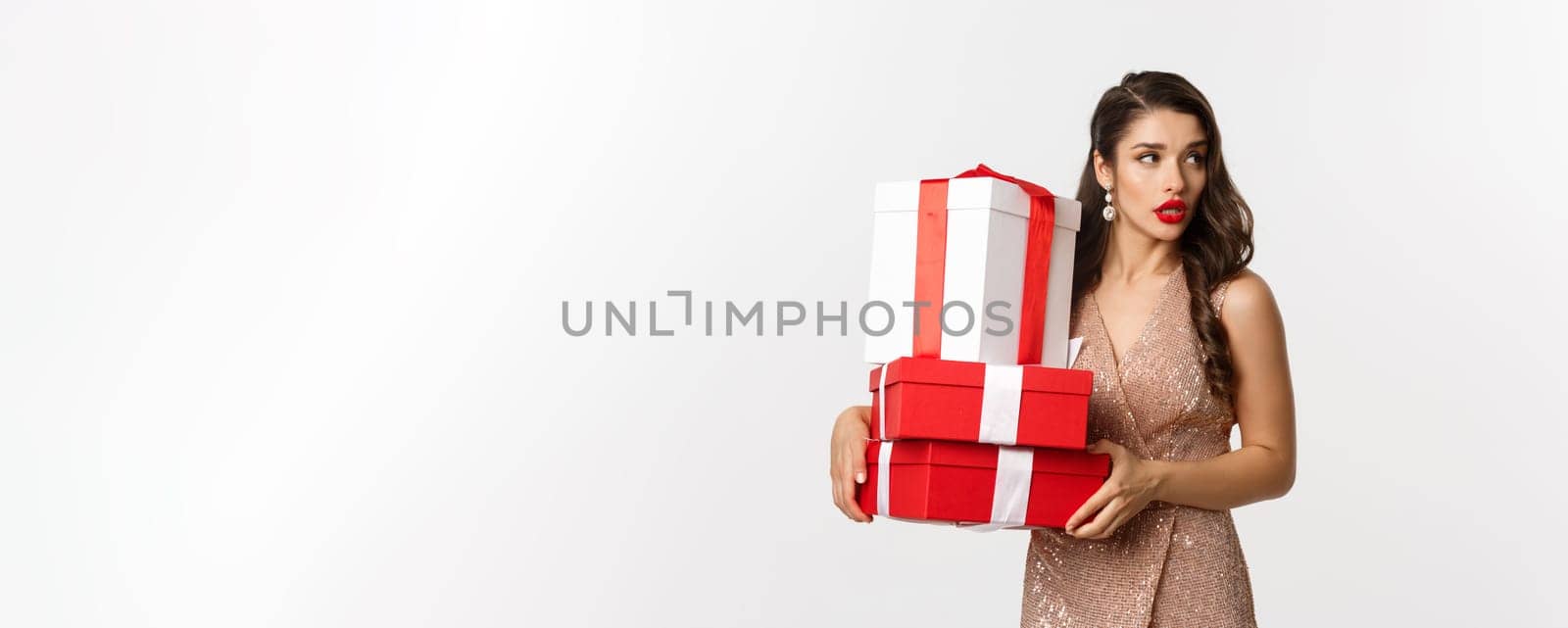 New Year, Christmas and celebration concept. Attractive young woman in elegant dress, looking away, carry gifts, standing over white background.