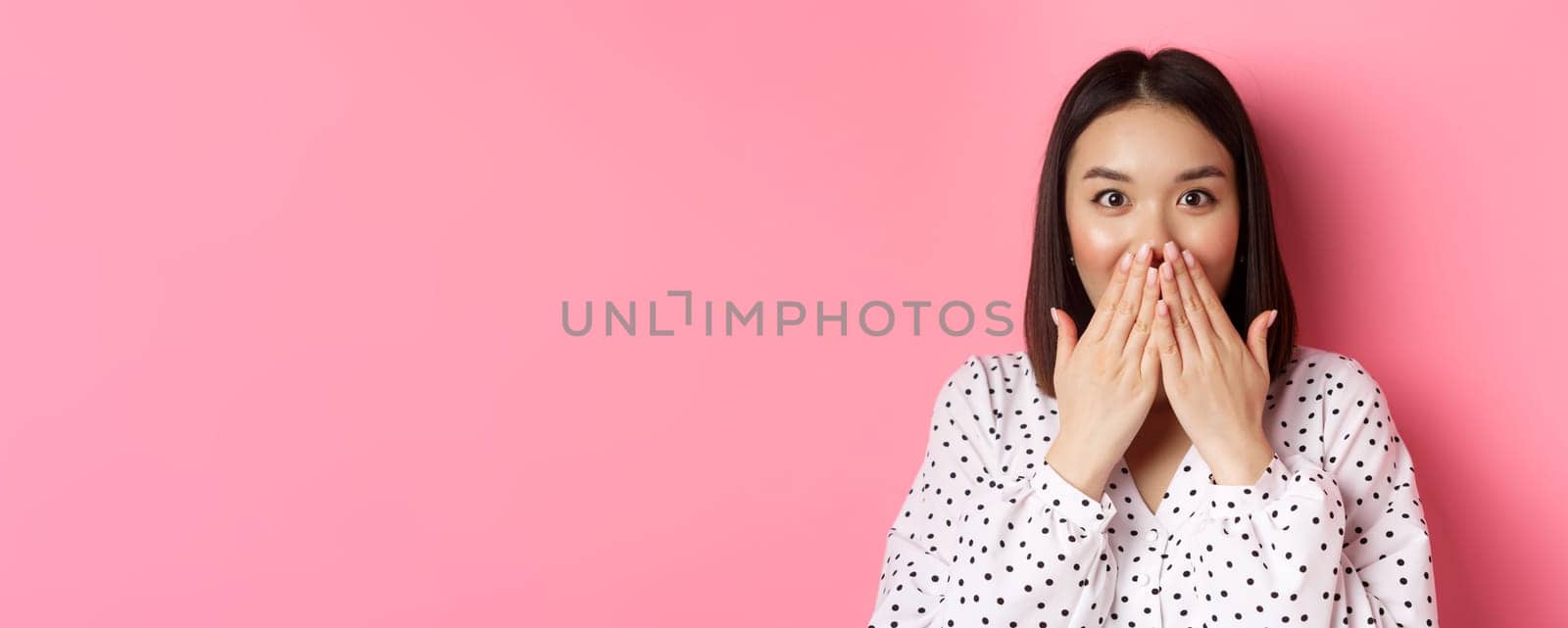Close-up of beautiful asian girl looking surprised and excited, cover mouth and staring amazed at camera, standing over pink background.