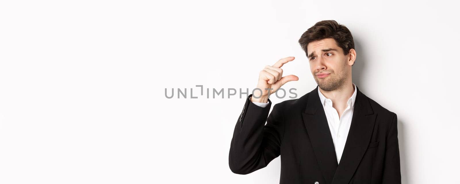 Close-up of handsome businessman in trendy suit, showing something small with disappointment, standing against white background.