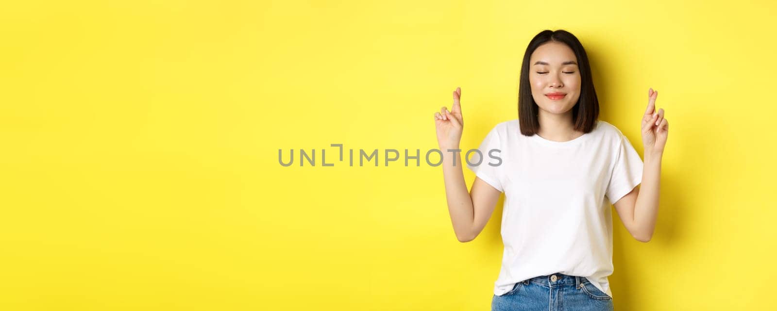 Hopeful asian girl making wish, cross fingers for good luck and praying with eyes closed, saying please, standing over yellow background.