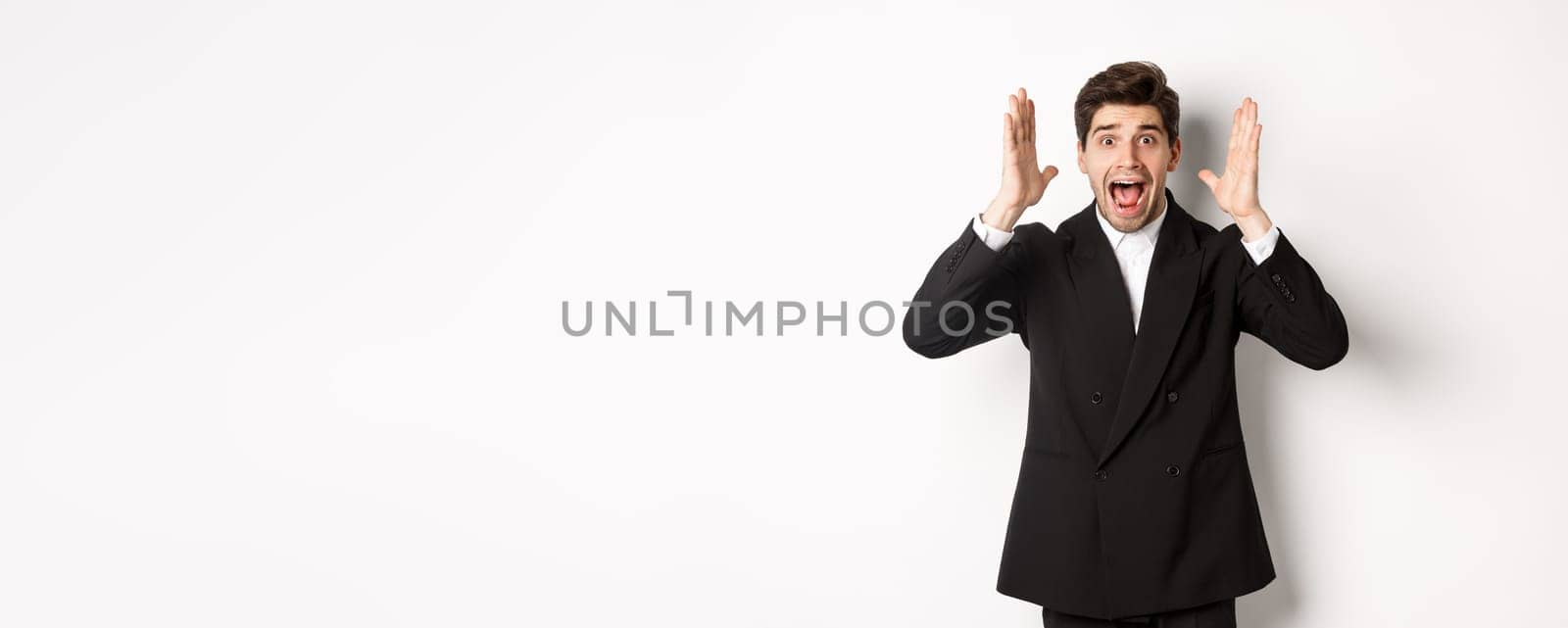 Frustrated and concerned man in black suit, screaming in panic and looking at something shocking, standing over white background.