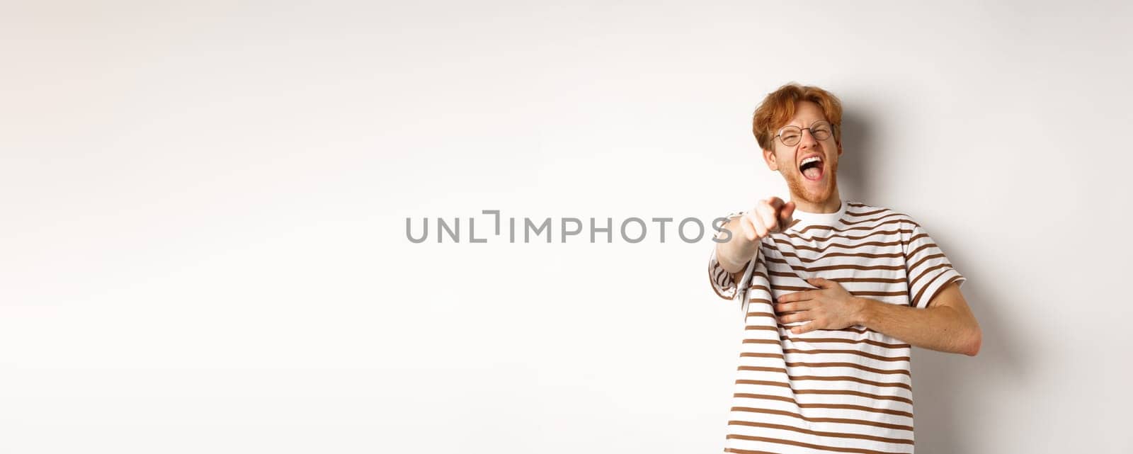 Young man with ginger hair and beard pointing finger at camera and laughing, making fun of someone hilarious, standing over white background.