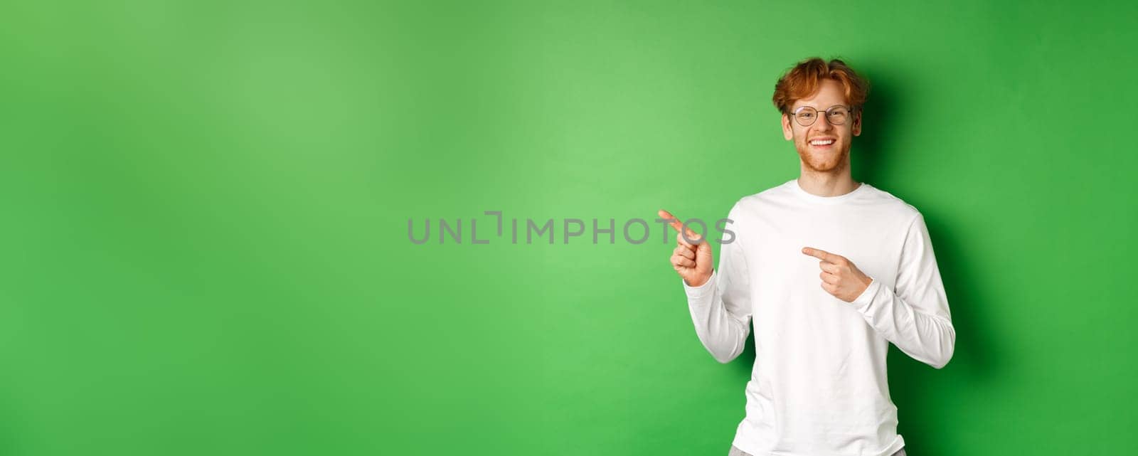 Handsome young man with red hair and beard smiling, pointing fingers left at logo, showing advertisement, standing over green background.