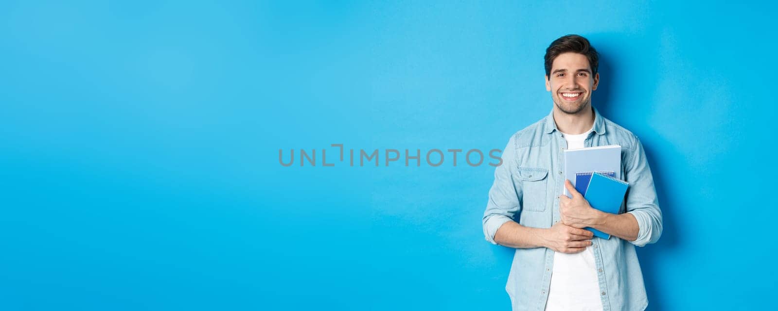 Young man holding notebooks and study material, smiling happy, standing over blue background.