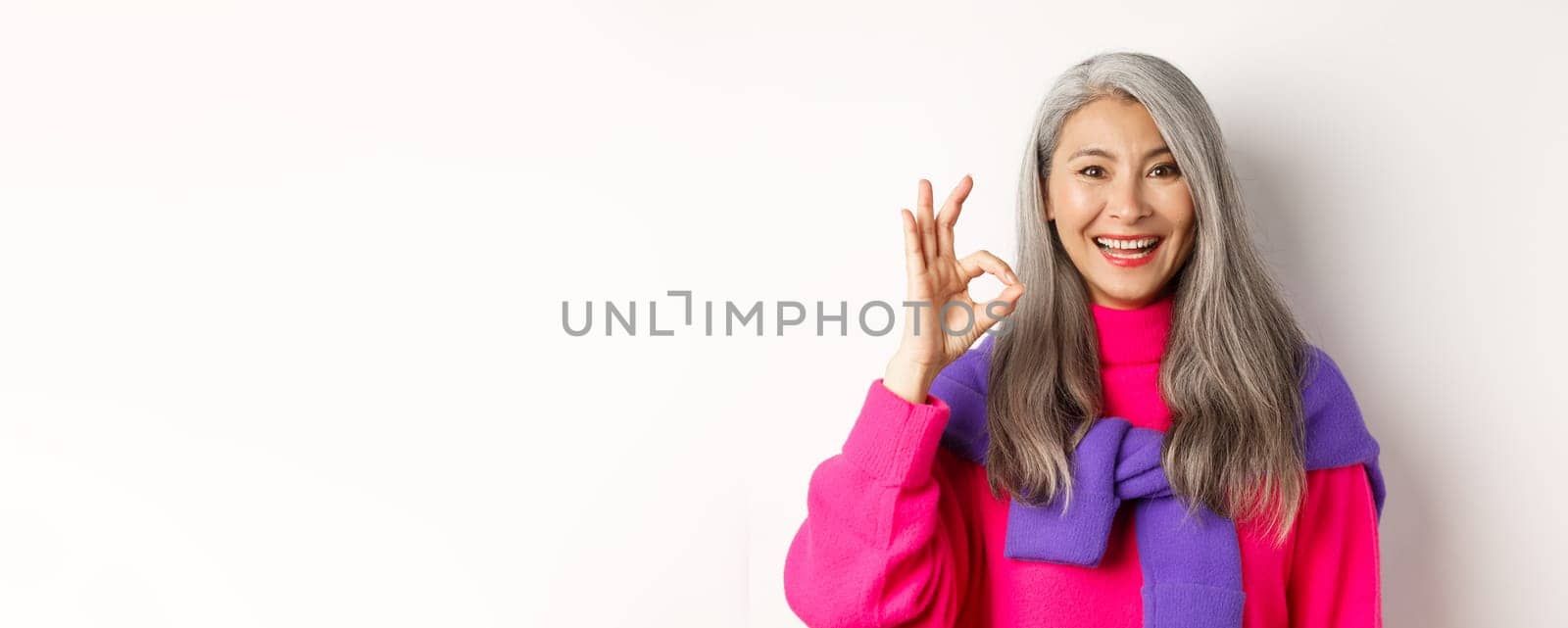 Close up of beautiful asian mature woman showing OK sign and smiling, approve and like product, standing satisfied against white background.